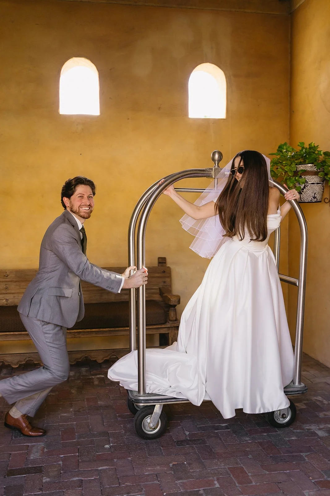 A man in a gray suit pulling a bride on a luggage cart inside a warmly lit room with yellow walls and arched windows.