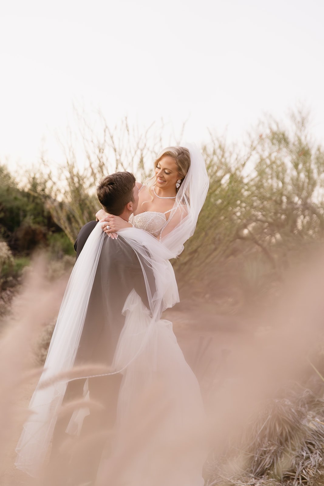 A bride and groom in wedding attire, outdoors among desert plants, with the groom lifting the bride in a loving embrace, she is smiling and wearing a veil and jewelry.