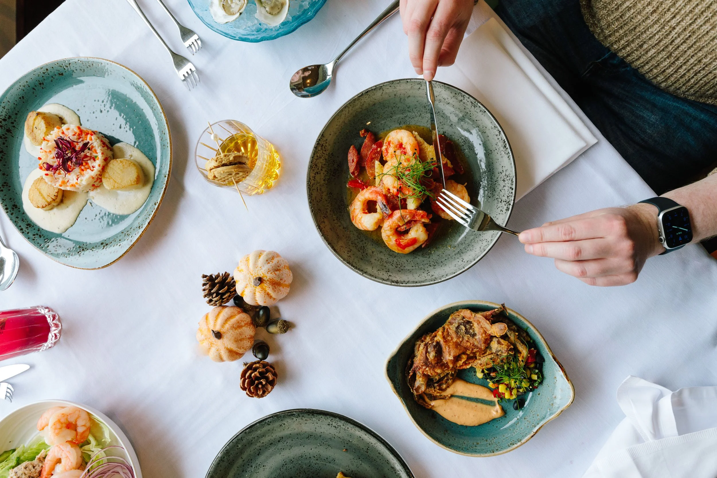Top view of a table set with various gourmet dishes including shrimp and sausage, scallops with puree, and a meat dish with sauce. A person's hand is using a fork and knife. The table is decorated with small pumpkins and pine cones, with drinks and additional seafood dishes.