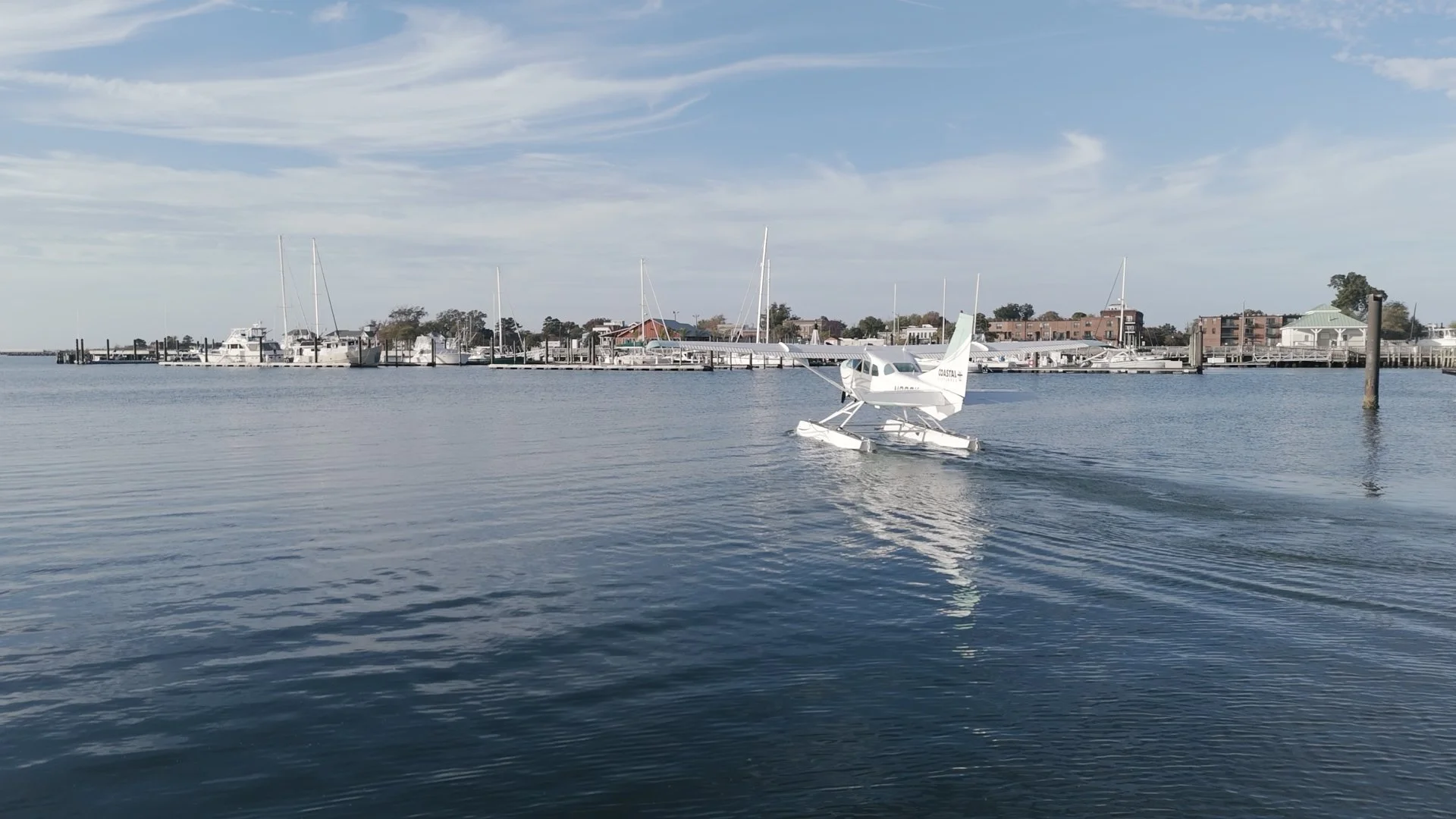 A seaplane on water near a marina with sailboats and buildings in the background, under a blue sky with clouds.