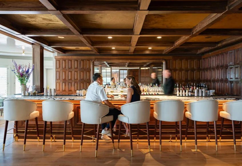 Elegant bar interior with wooden decor, couple seated at counter, bartender moving, flower arrangement