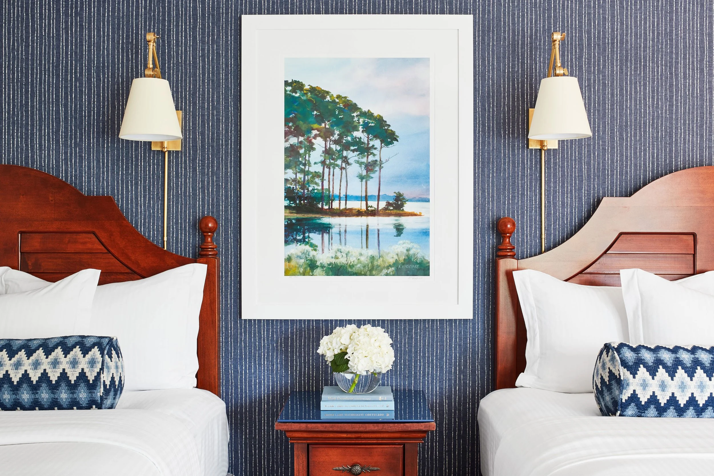 Hotel room with two wooden beds, blue pillows, floral artwork, wall lamps, and a nightstand with books and flowers.