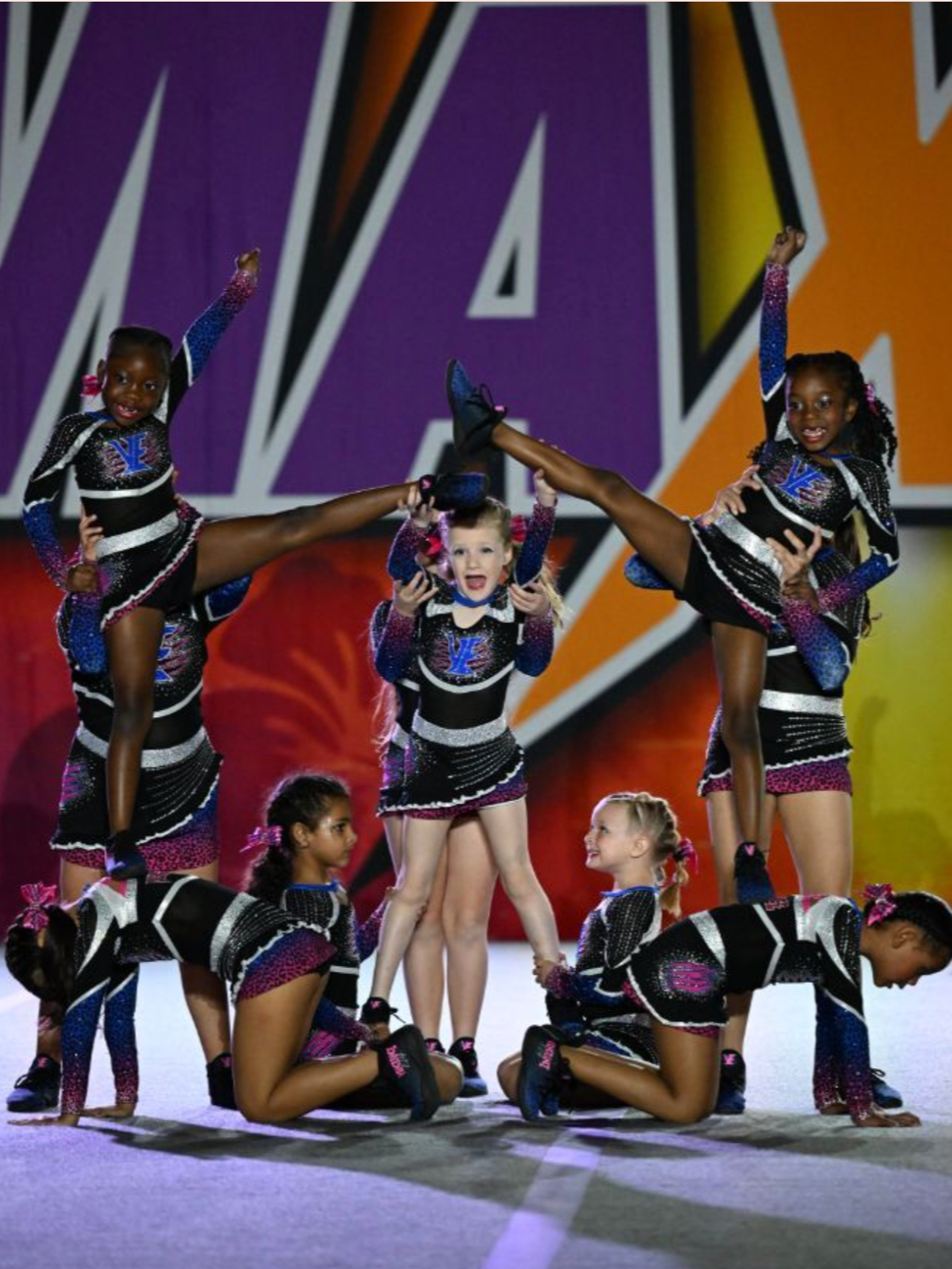 Young gymnasts in matching sequined leotards performing a formation on stage, with some standing, some kneeling, and two girls lifted in the air, against a bright, colorful background.