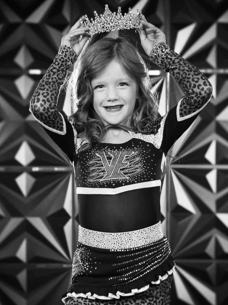 Young girl smiling and holding a tiara above her head, wearing a cheerleading uniform with leopard print sleeves and a sparkling waistband, standing in front of geometric background.