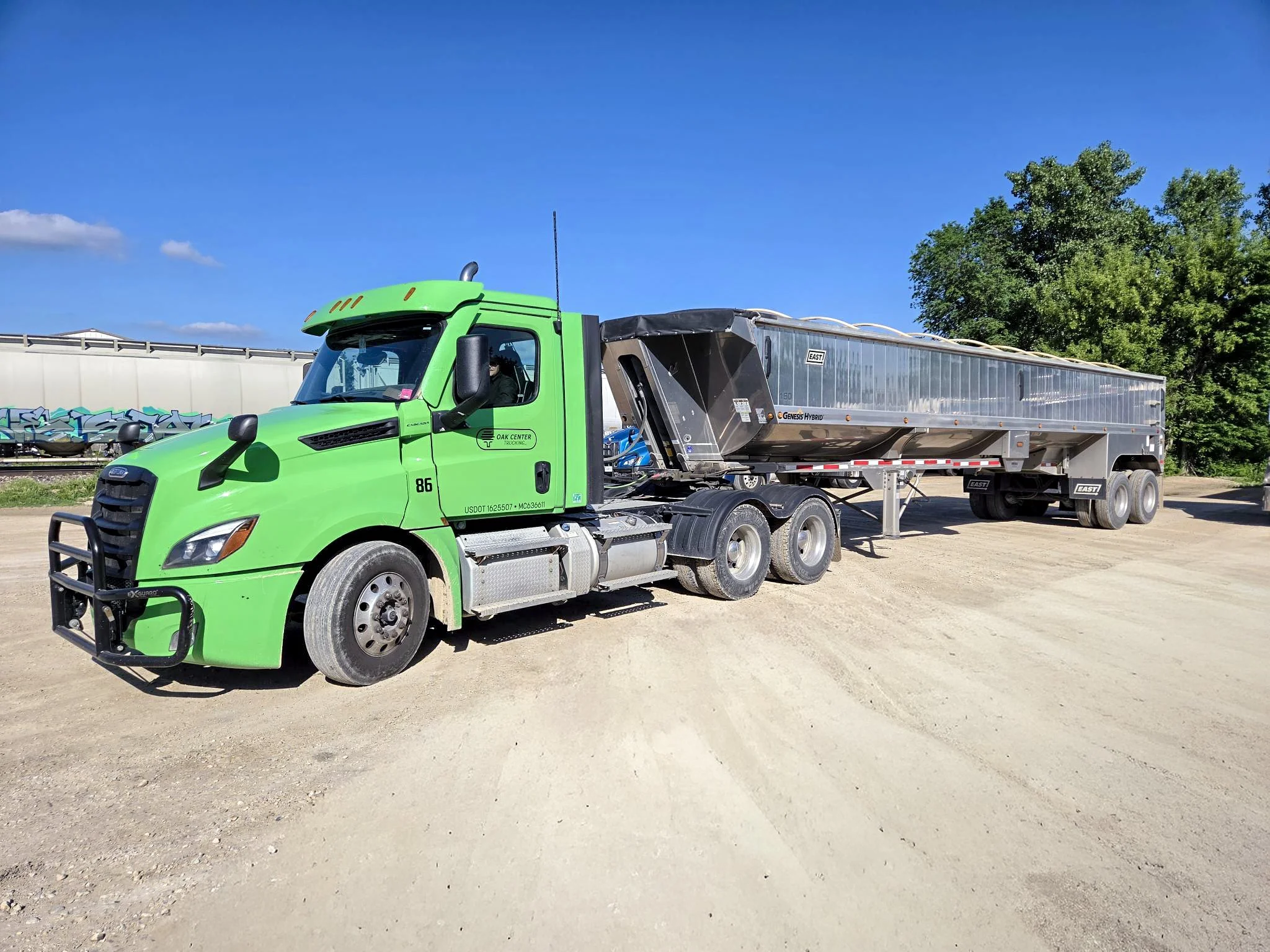 Green semi-truck with a silver trailer driving on a dirt road under a blue sky with some clouds, next to green trees.