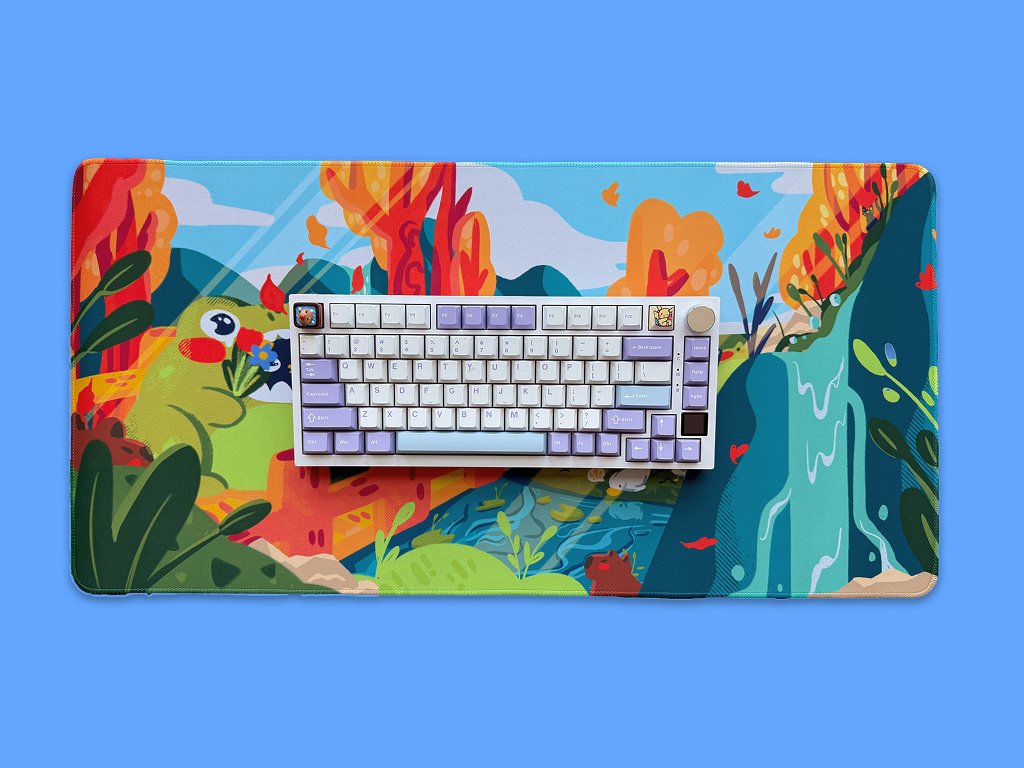 A colorful illustrated desk mat with a scenic nature landscape, featuring mountains, trees with autumn foliage, a waterfall, and a frog holding a flower, placed on a blue background. A compact mechanical keyboard with pastel purple, white, and light 