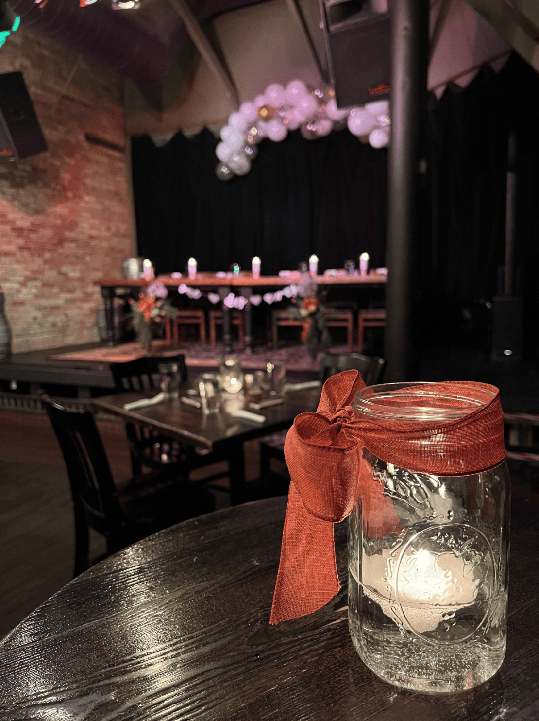 A decorated event space with a mason jar candle holder wrapped with a red ribbon on a wooden table, set for dining and a stage with floral and balloon decorations in the background.