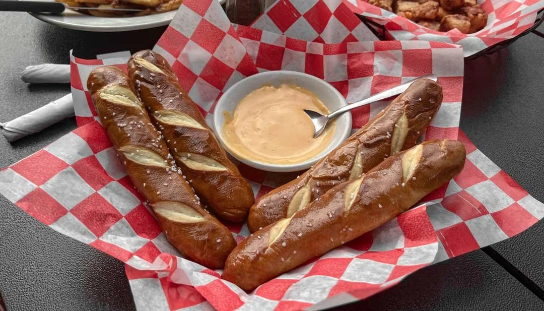 Three breadsticks with salt, served with a bowl of creamy dipping sauce, on red and white checkered paper in a basket.