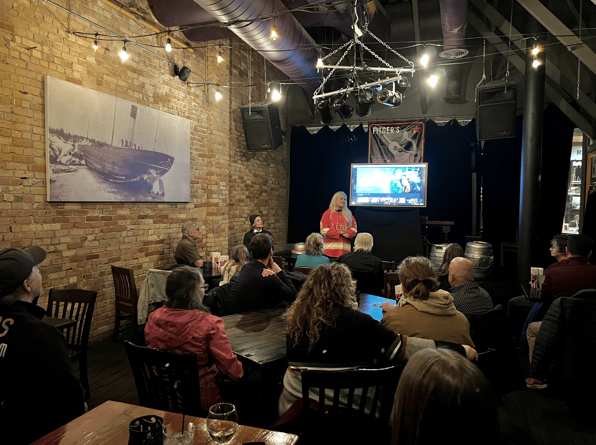 A group of people watching a woman speaking on stage in a dimly lit room with brick walls, a large screen, and hanging string lights.