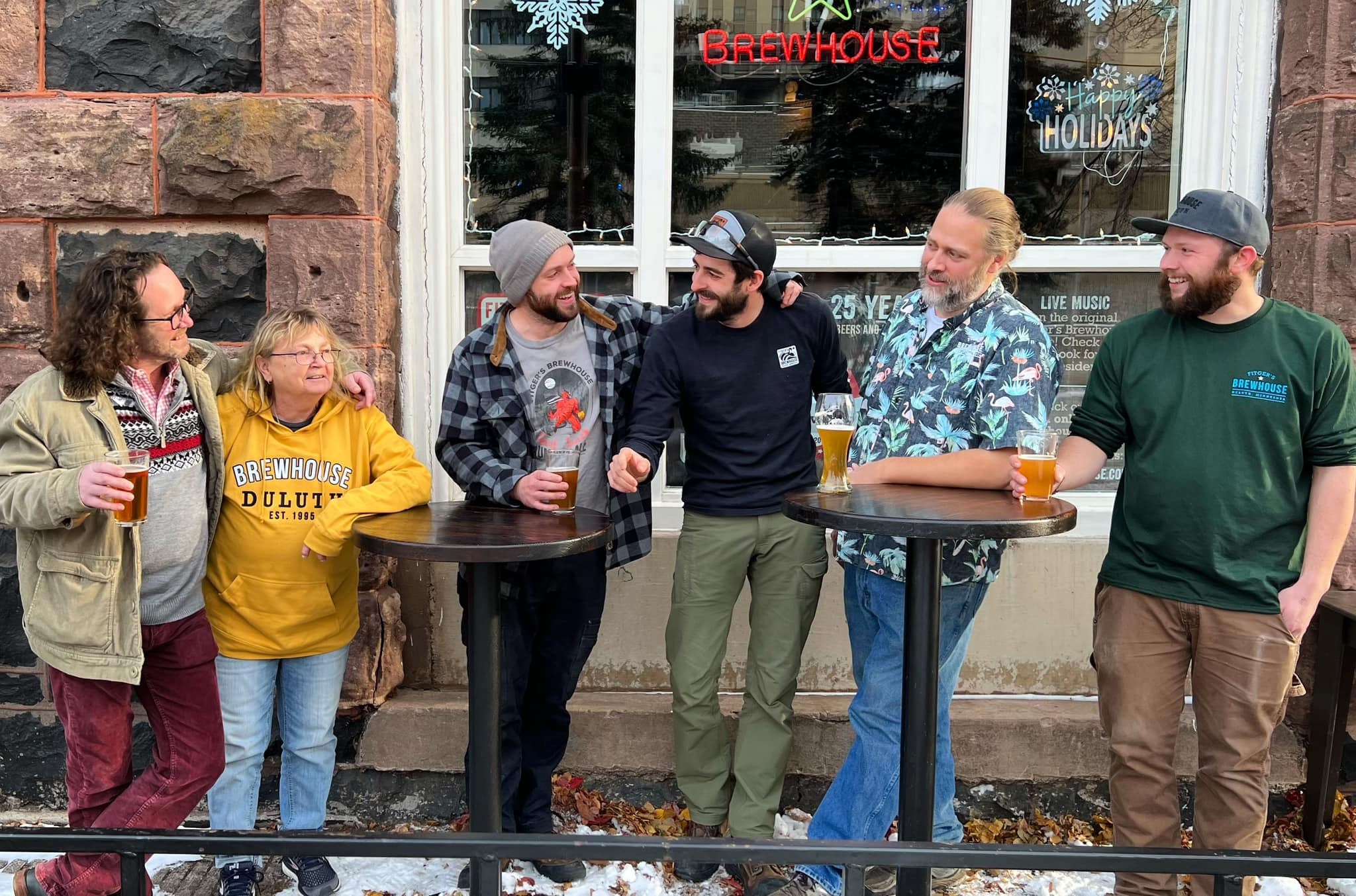 Six people standing outside at high tables with drinks, chatting and laughing in front of a brewhouse window decorated with holiday signs and neon lights.