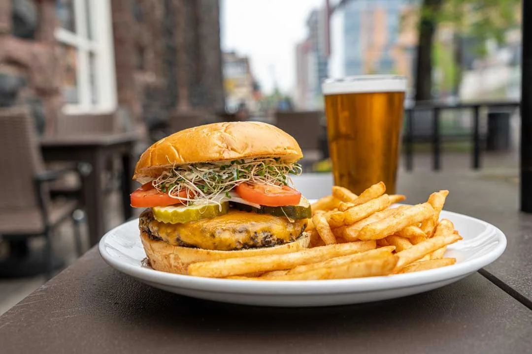 Plate with cheeseburger, French fries, and a glass of beer on an outdoor table in an urban setting.