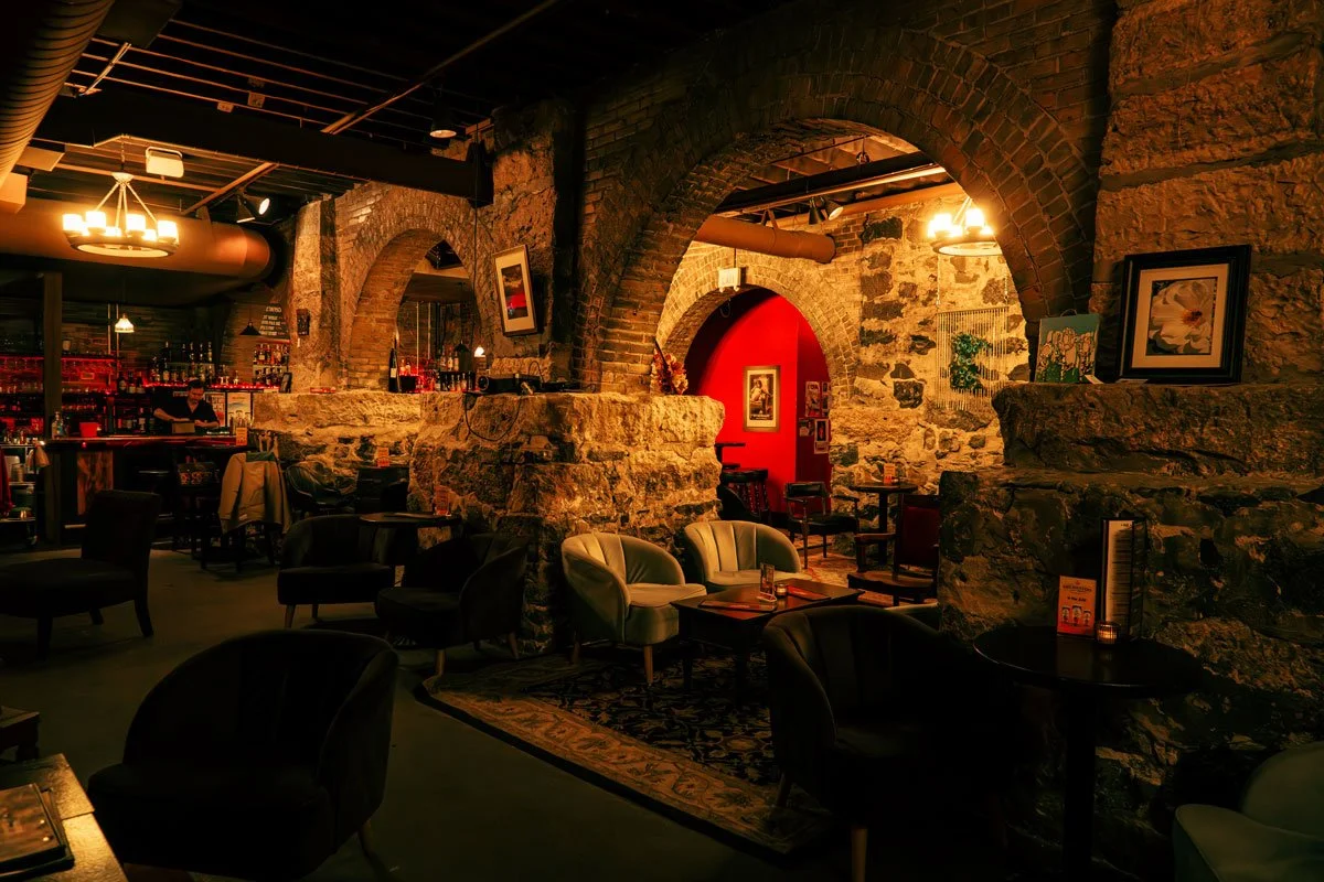Cozy bar or lounge with stone arches and walls, dim lighting, and comfortable chairs and tables.