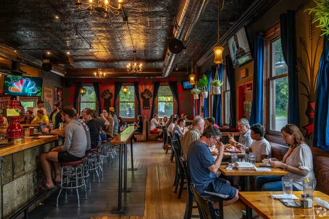 A lively restaurant with a bar and guests dining, decorated with artwork of raised fists on the walls, blue curtains, and hanging plants.