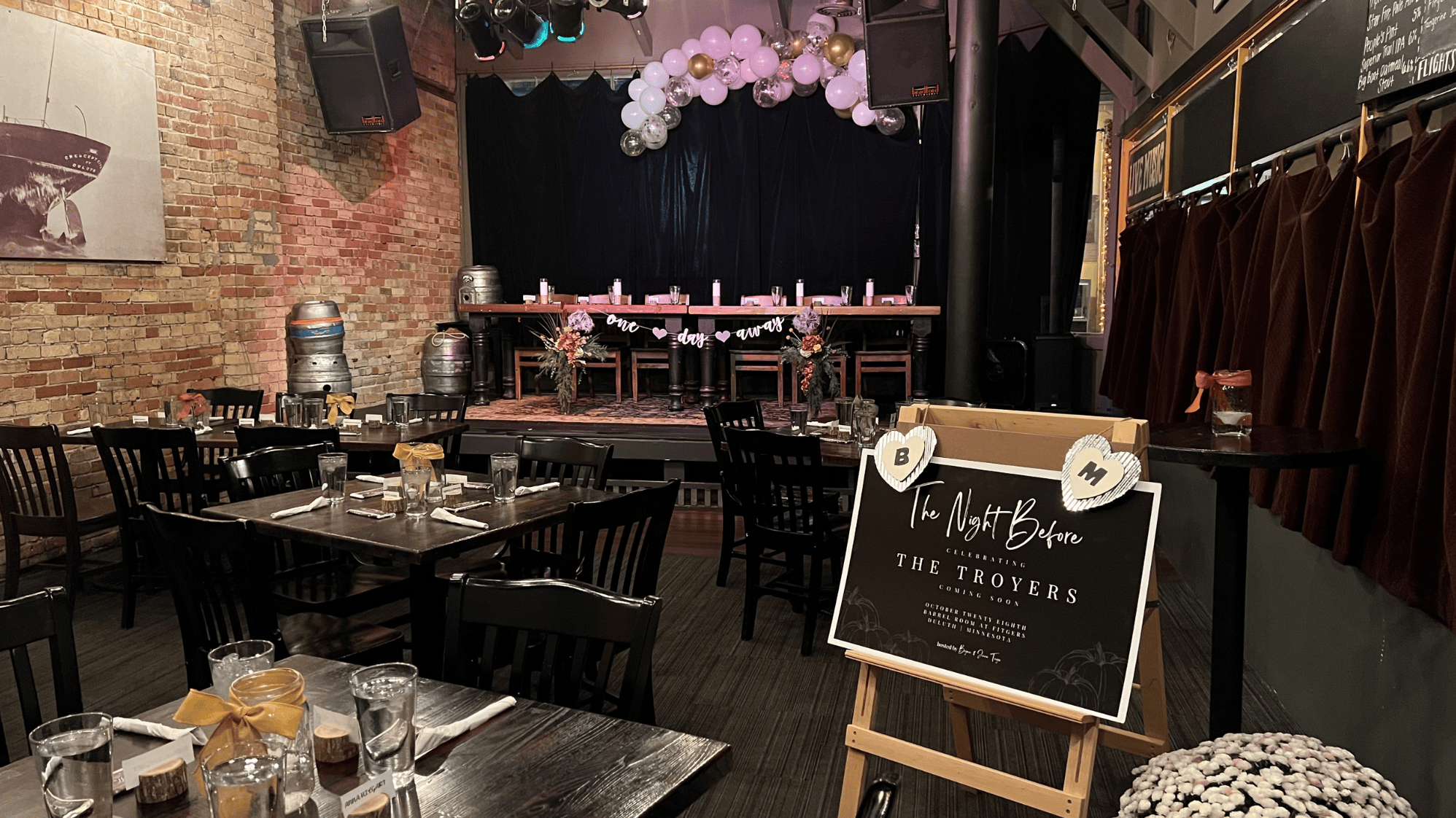 An indoor venue set for a celebration with a stage decorated with balloons, flowers, and a banner, and tables arranged with glasses, napkins, and decorative bows, preparing for an event called 'The Night Before' featuring 'The TROYERS'.