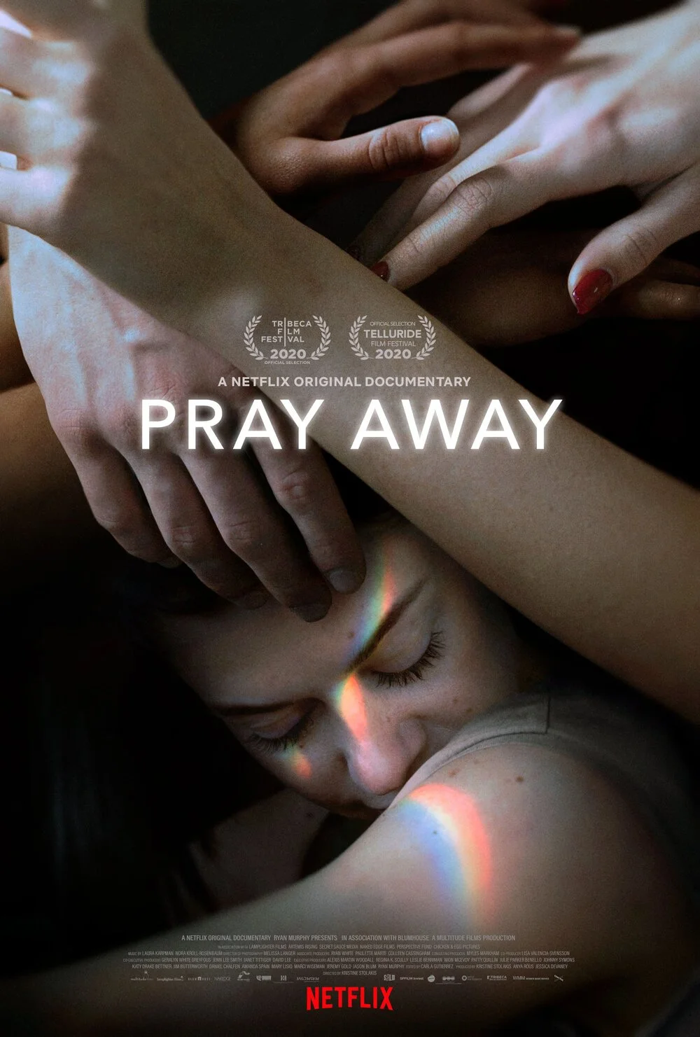 Pray Away film poster with photograph of multiple hands crisscrossing to touch each other, with one hand touching the forehead of a white person with freckles and closed eyes, the reflection of a rainbow shines over their face and upper arm.