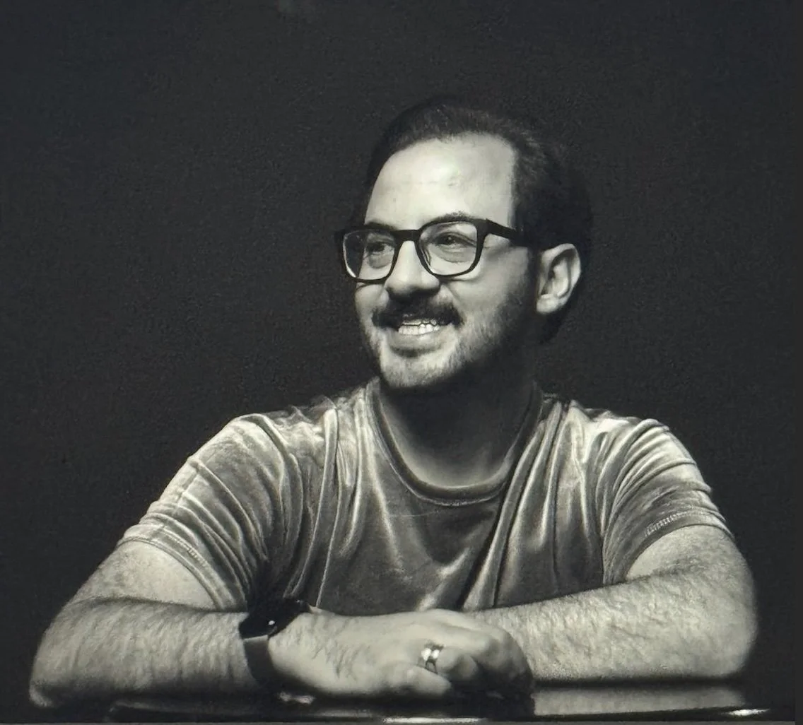 Black and white headshot of Felipe Estafan. Felipe is a Latino man with a moustache, square black glasses and thick short hair. He looks to the left, smiling in a photo studio.