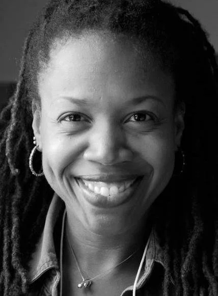 Black and white headshot of Natalie Bullock Brown. She is a Black woman with small hoop earrings, pulled back locs and a wide smile as she looks into the camera.