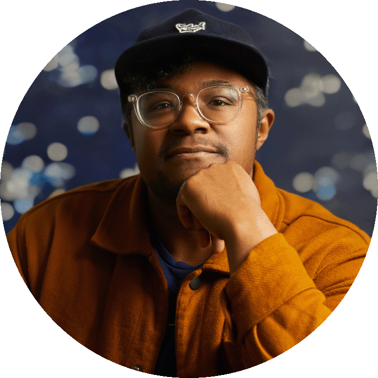 Brit is a Black trans person with thin-rimmed glasses looking off to the left. He is wearing a black shirt with a grey outer layer against a brown backdrop.