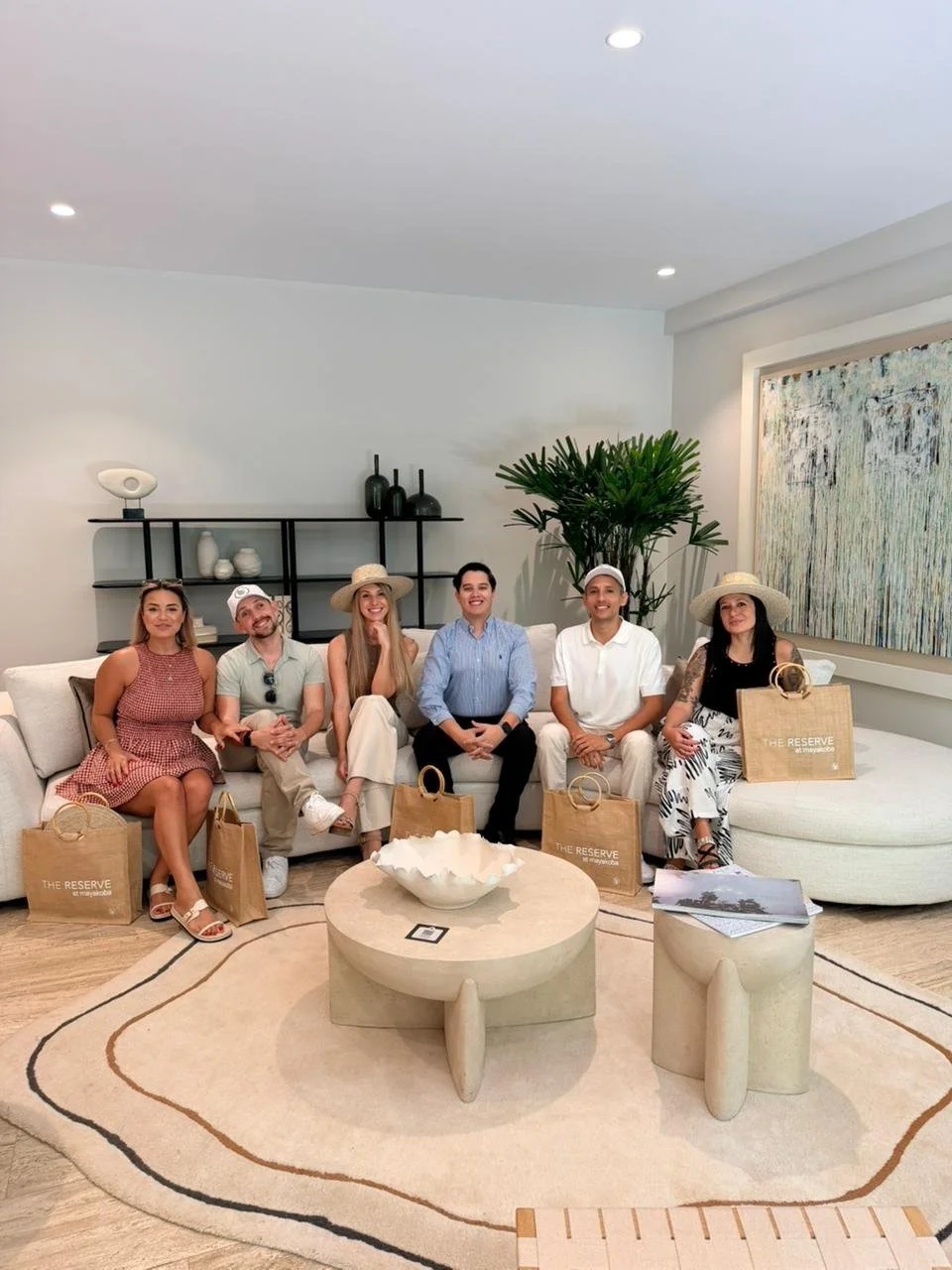 Six people sitting on a white sofa in a modern living room, with shopping bags labeled 'The Reserve' placed next to them, and a large abstract painting on the wall behind. They appear to be celebrating or attending an event.