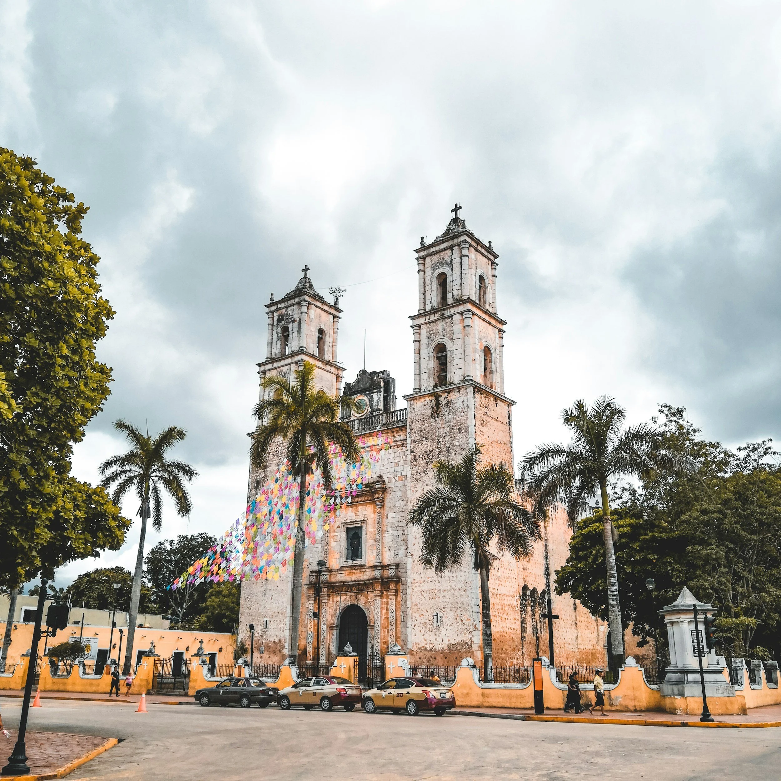 A historic church with twin bell towers decorated with crosses, surrounded by palm trees, with a cobblestone street in front and parked cars, under a cloudy sky.