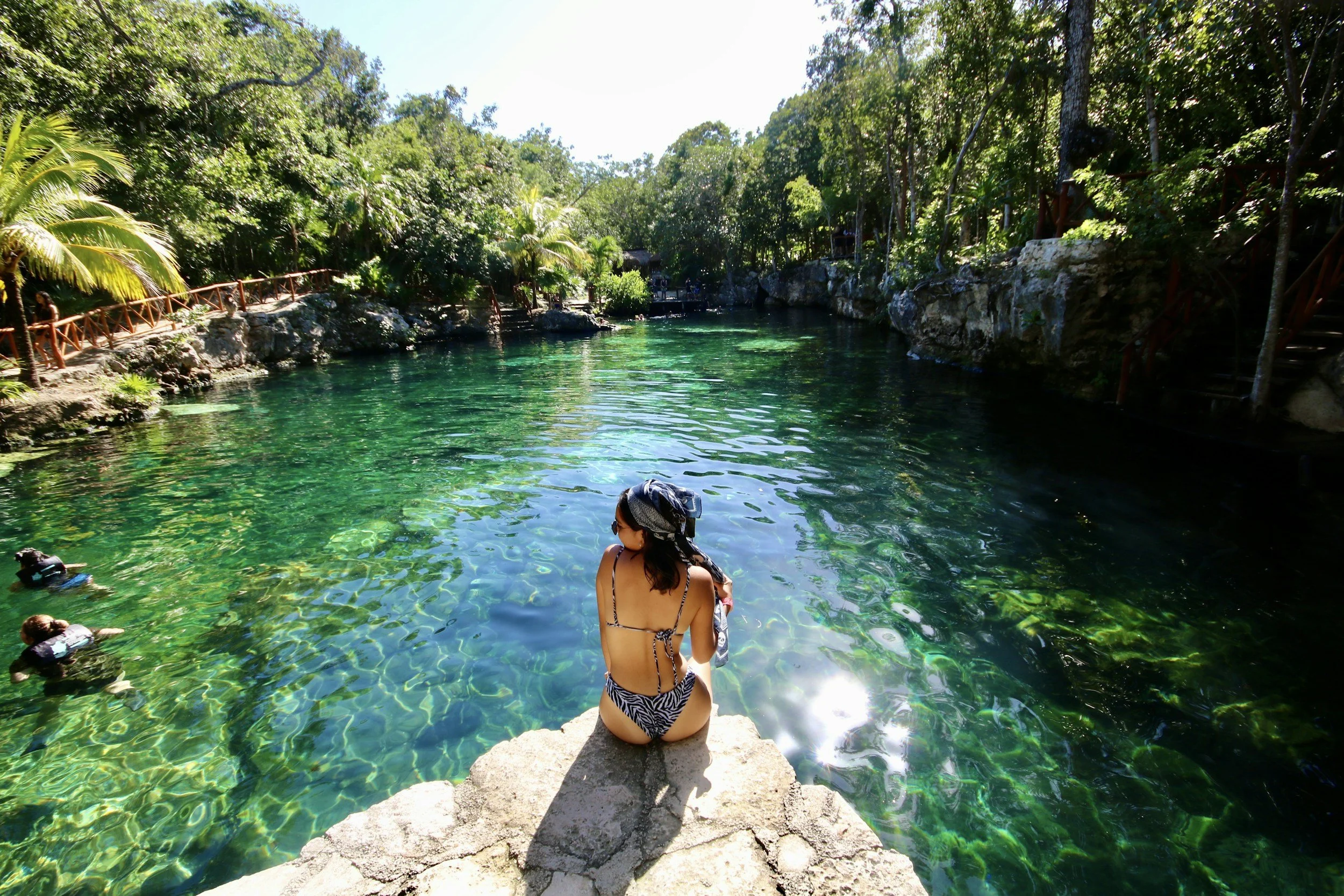 A woman in a swimsuit with a bandana sitting on a rock at the edge of a clear, green water cenote surrounded by lush trees.