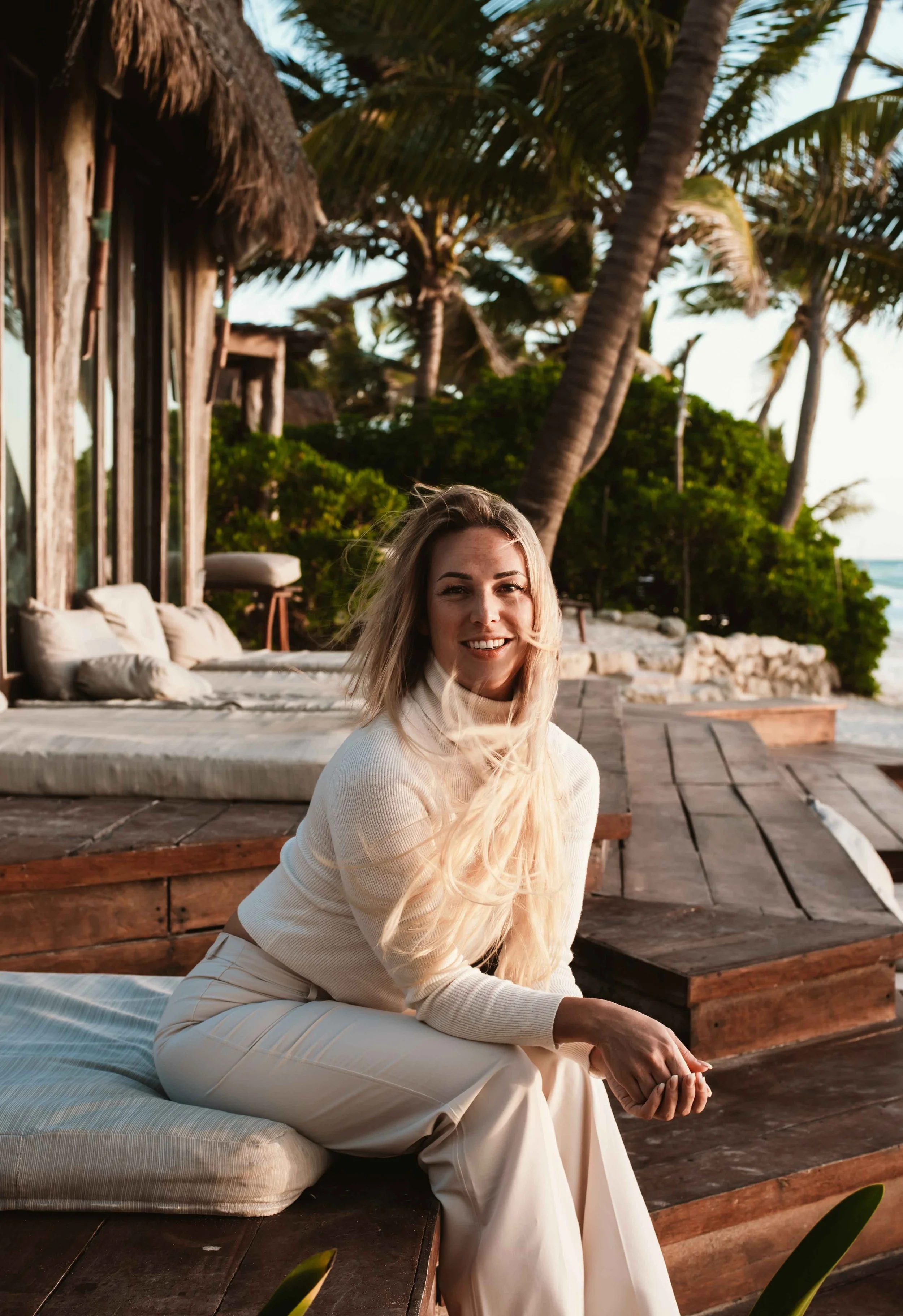 A woman with long blonde hair and a cream-colored sweater seated on a cushioned bench on a wooden deck with tropical trees and the ocean in the background.