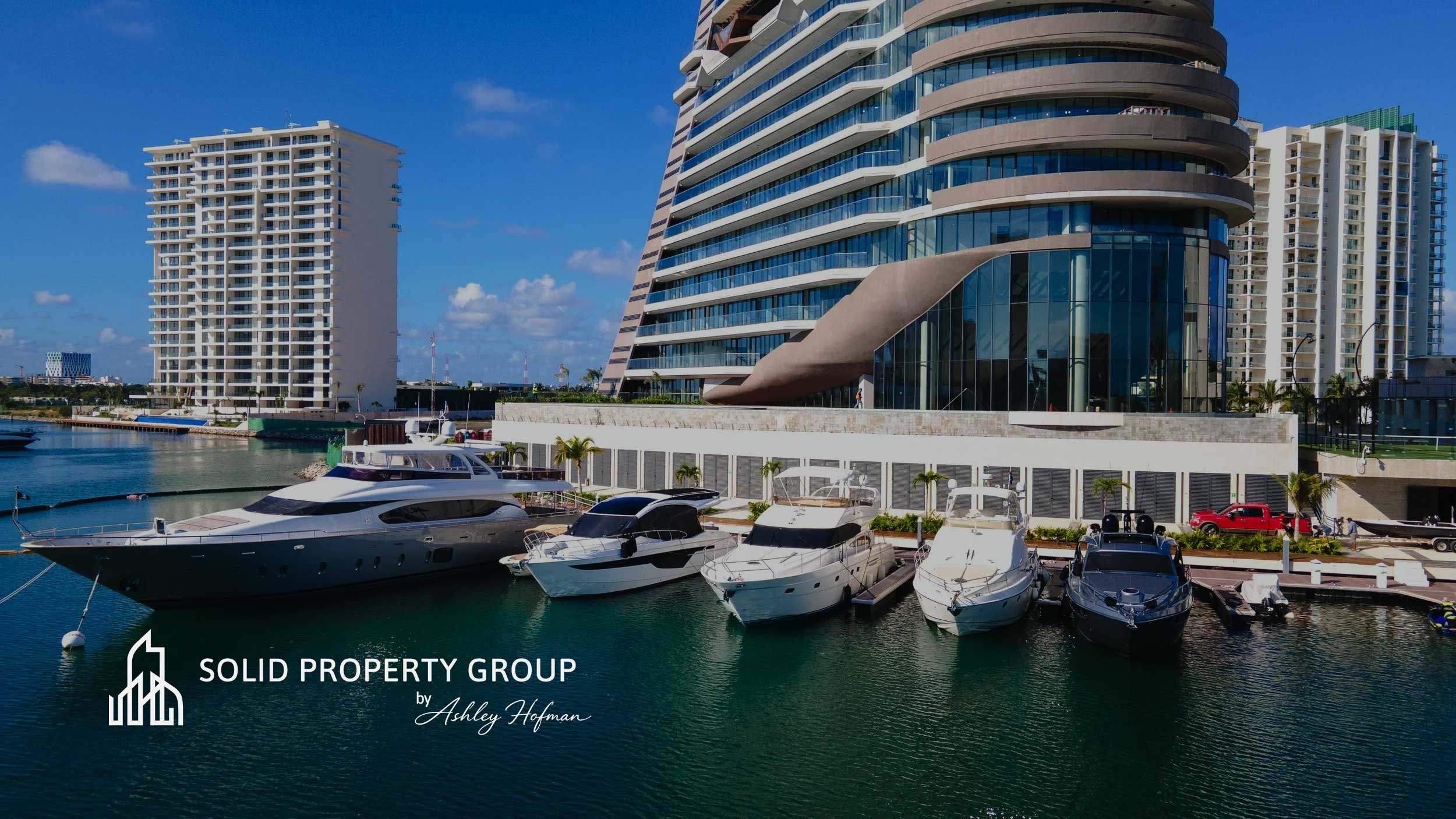 Luxury boats docked at a marina with modern high-rise buildings in the background under a blue sky.