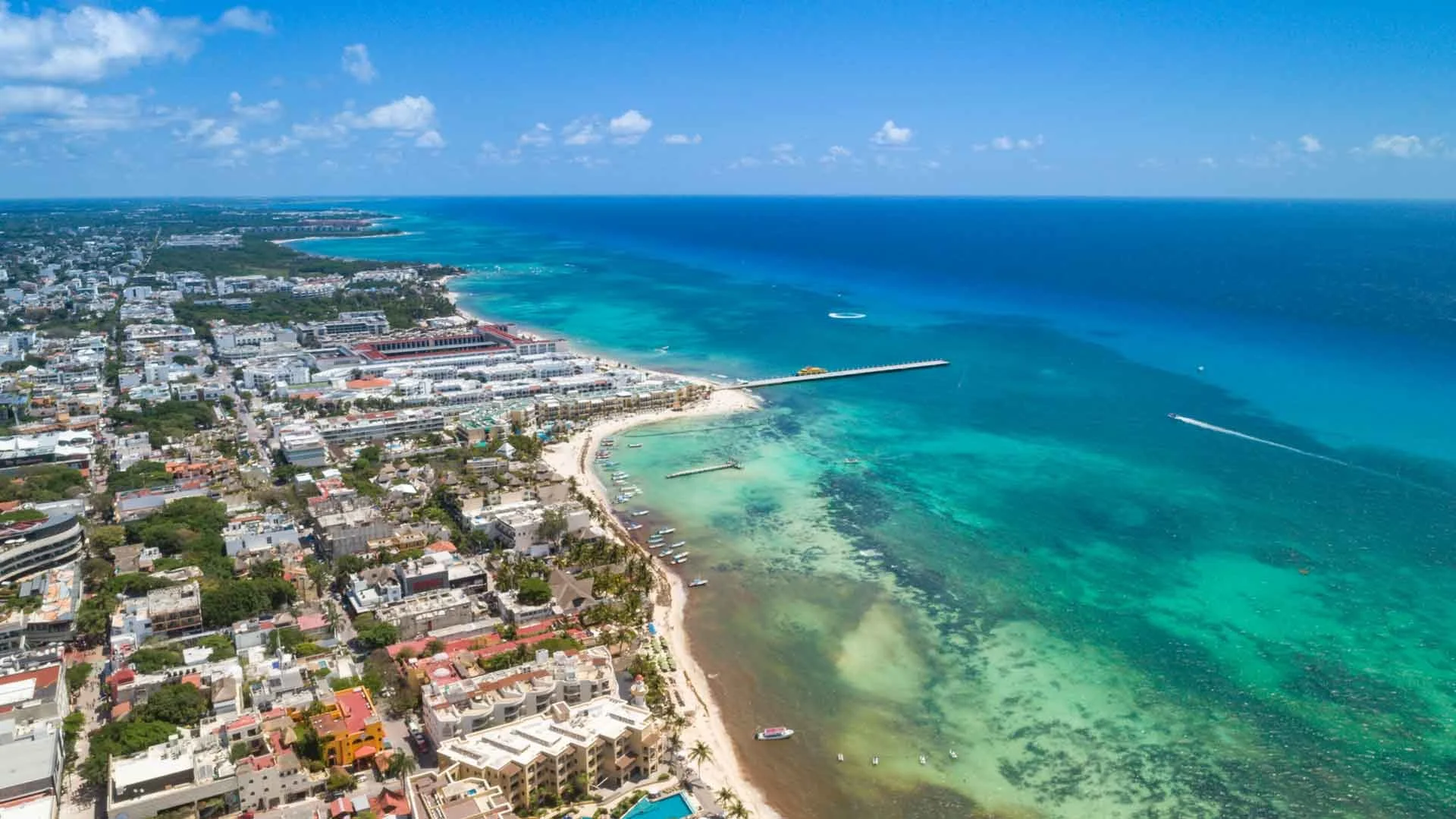 Aerial view of a coastal city with turquoise water, sandy beaches, high-rise buildings, and a breakwater extending into the ocean under a blue sky with some clouds.