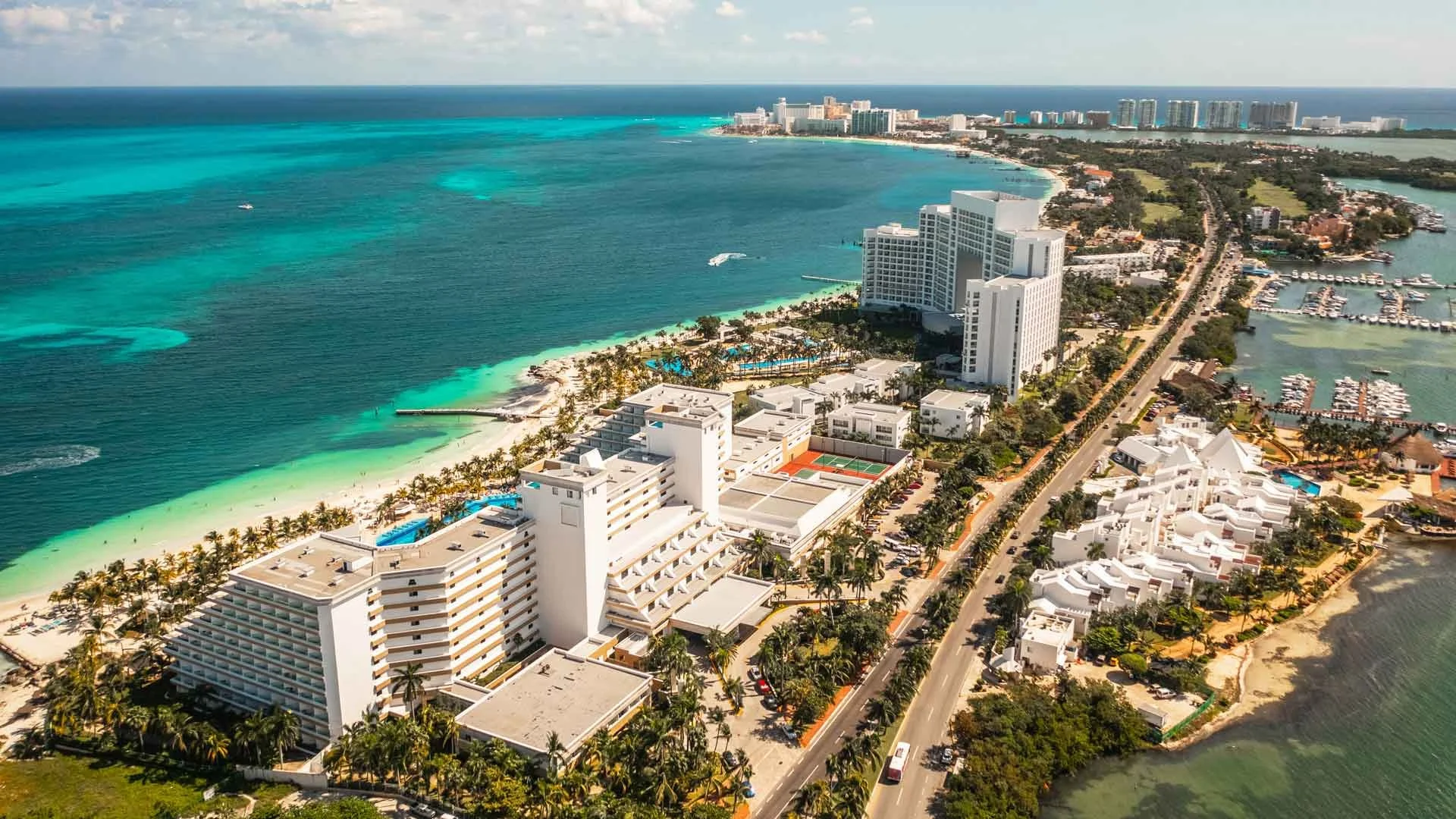 Aerial view of a coastal city with high-rise hotels, white residential buildings, palm trees, a beach with turquoise water, a marina with boats, and a main road running parallel to the shoreline.