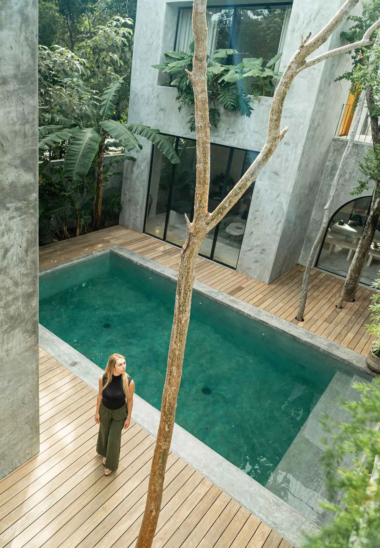 A woman walking on a wooden deck beside a rectangular swimming pool with green water. The background features a modern concrete building with large glass windows and lush green foliage and trees.