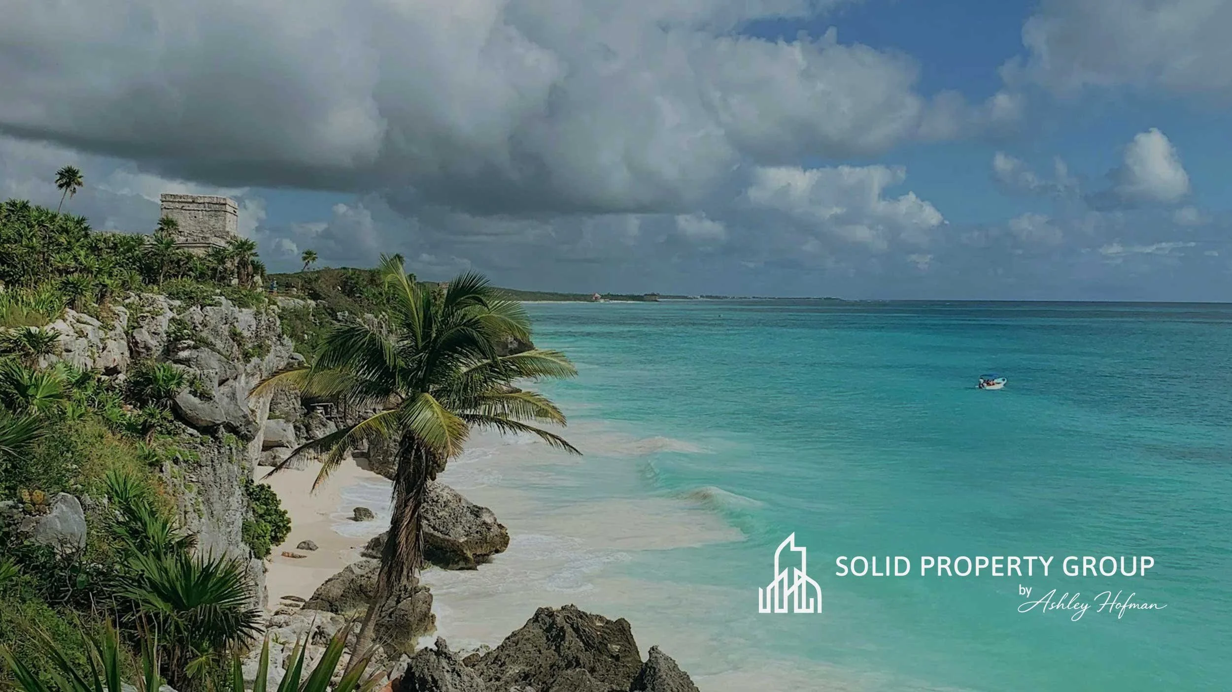Tropical beach with turquoise water, white sandy shore, palm trees, and rocky cliffs under a partly cloudy sky. There is a small boat in the water and a logo for Solid Property Group by Ashley Hofman in the bottom right corner.