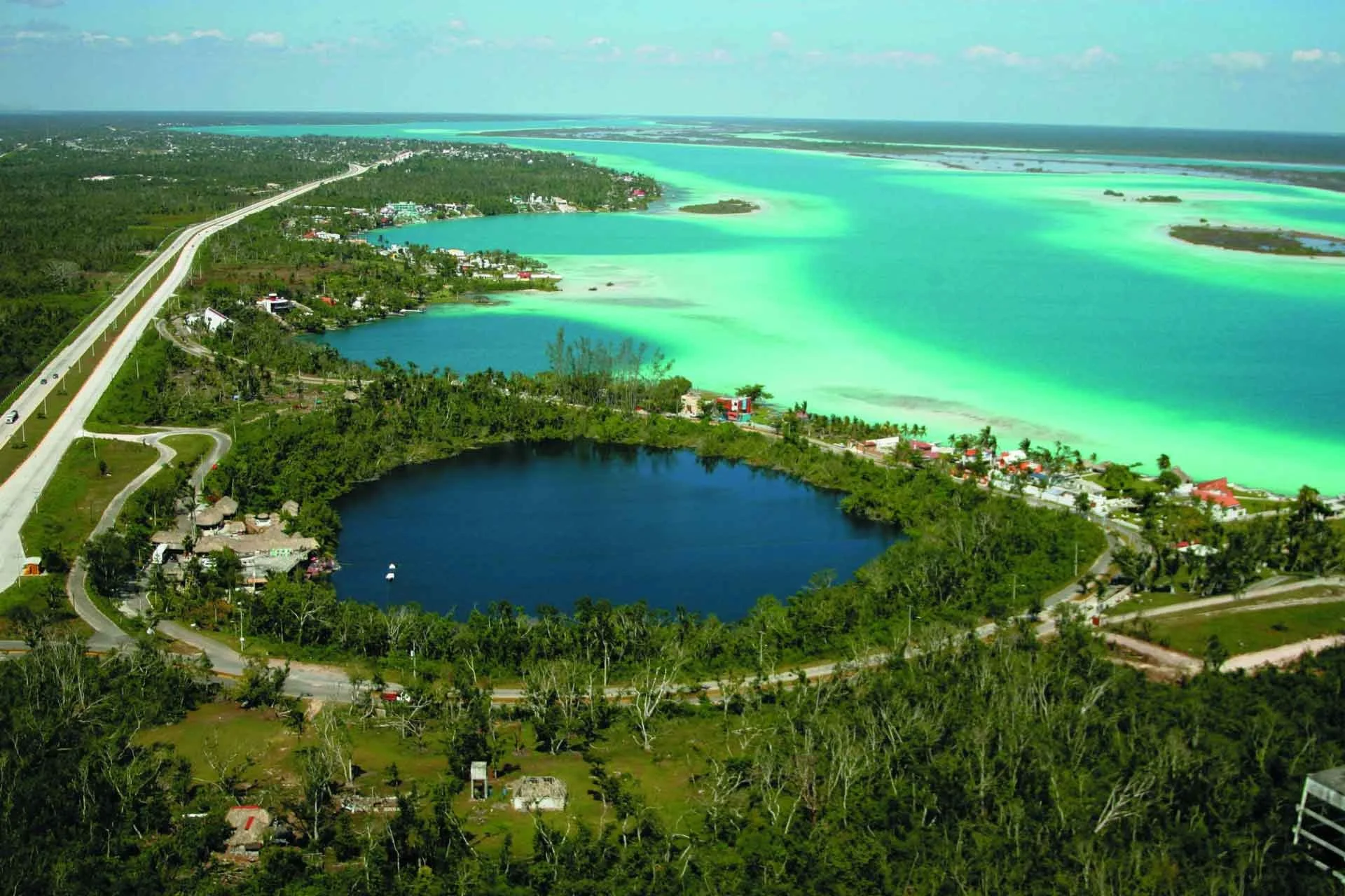 Aerial view of a coastal area with a large dark lake surrounded by green trees and houses, a bridge crossing over the landscape, and turquoise and light blue ocean waters in the background under a partly cloudy sky.