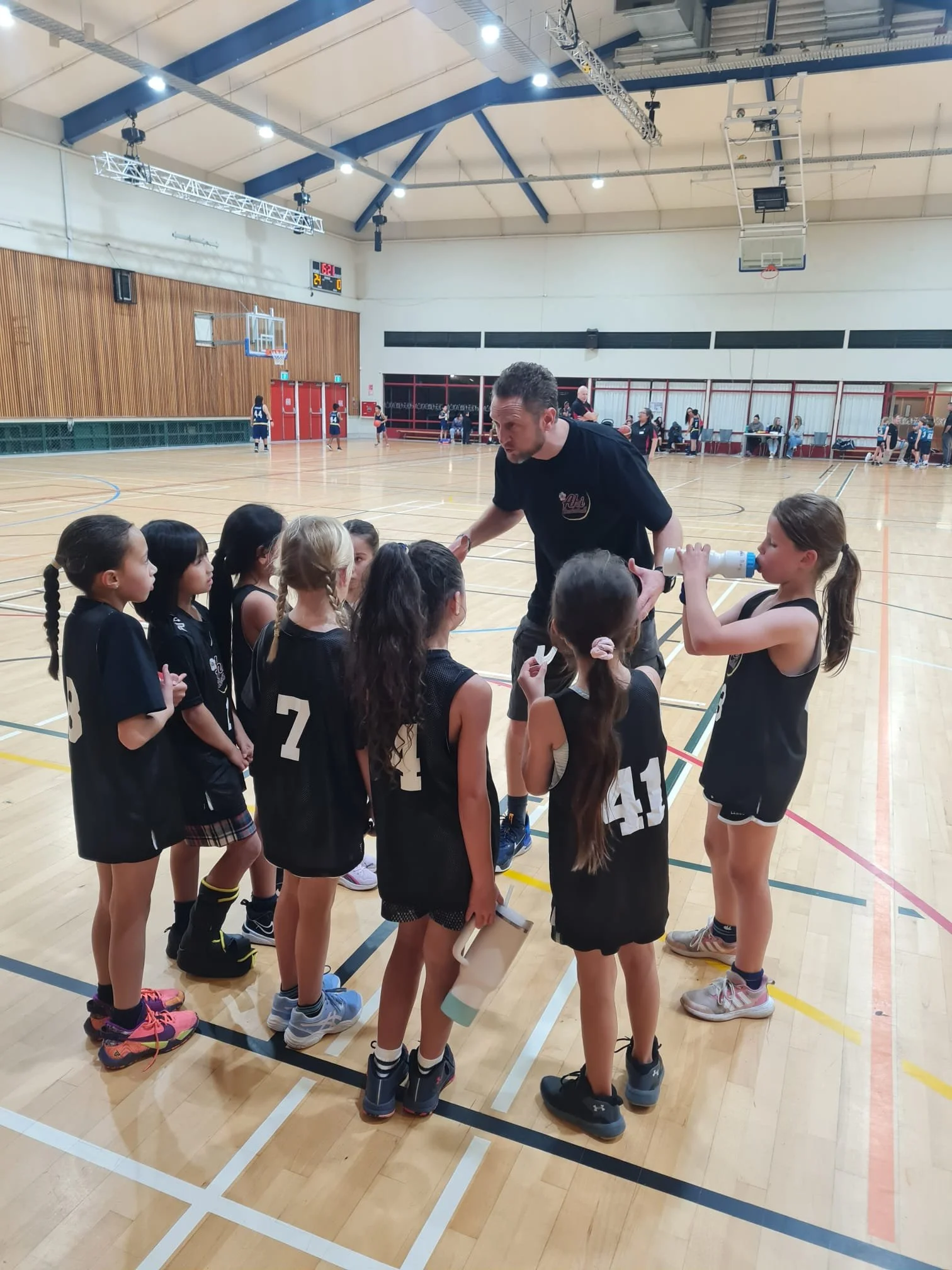 A girls' basketball team huddle in a gym, listening to their coach. The gym has a wooden floor, basketball hoops, and a scoreboard. Other players are practicing on the court in the background.