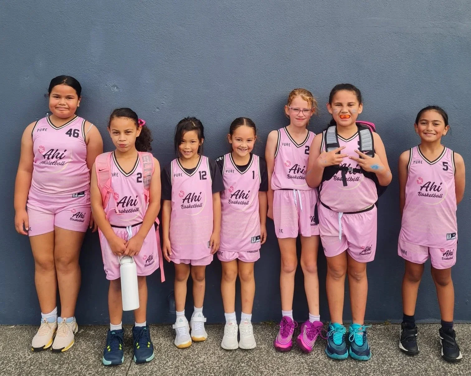Group of seven young girls in pink basketball uniforms standing against a blue wall.