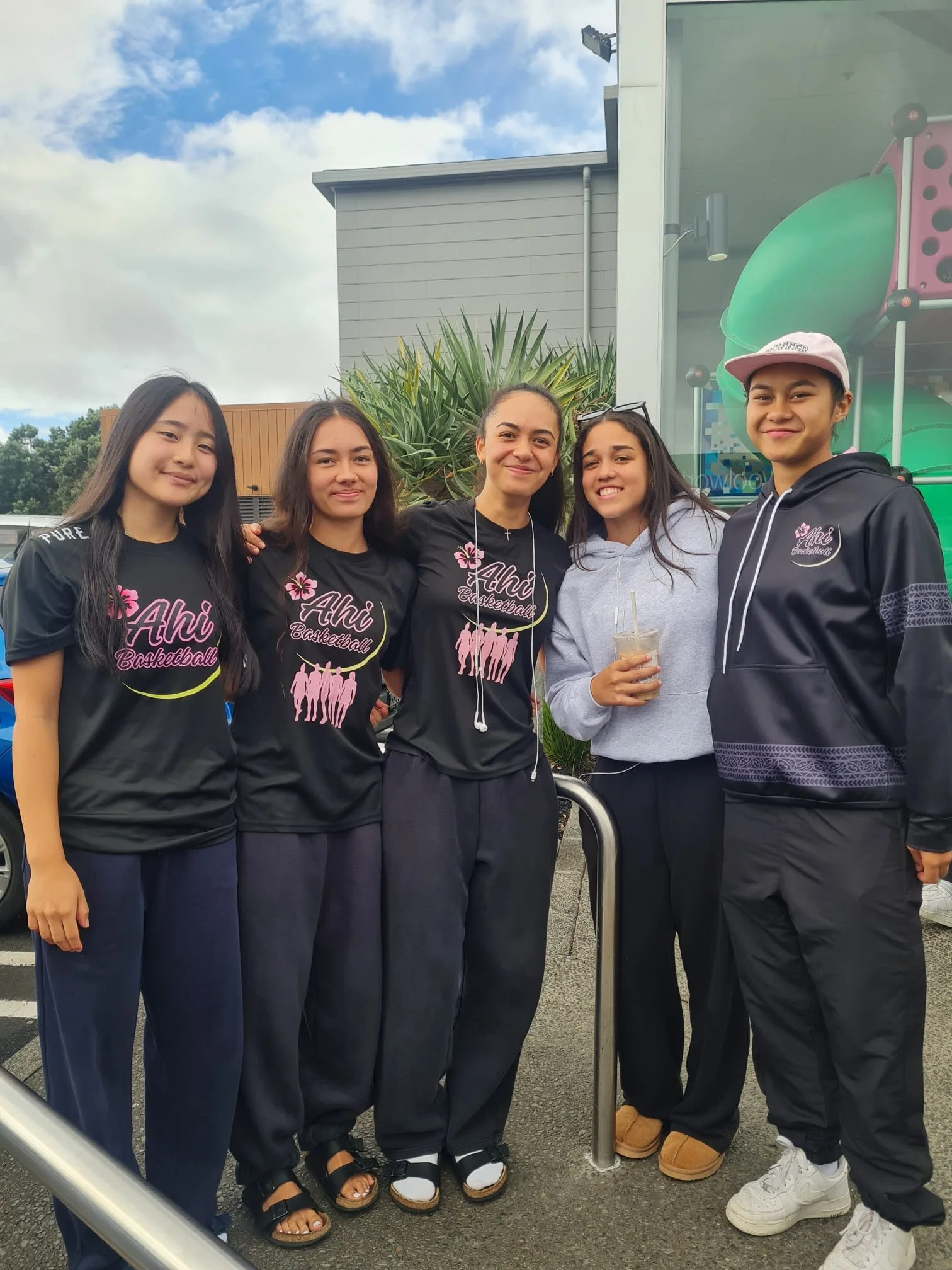 A group of five teenagers, four females and one male, standing together outdoors. The group appears happy, with some smiling and embracing. The background includes green plants, a building, and a blue sky with clouds. One girl is holding a drink in a clear plastic cup.