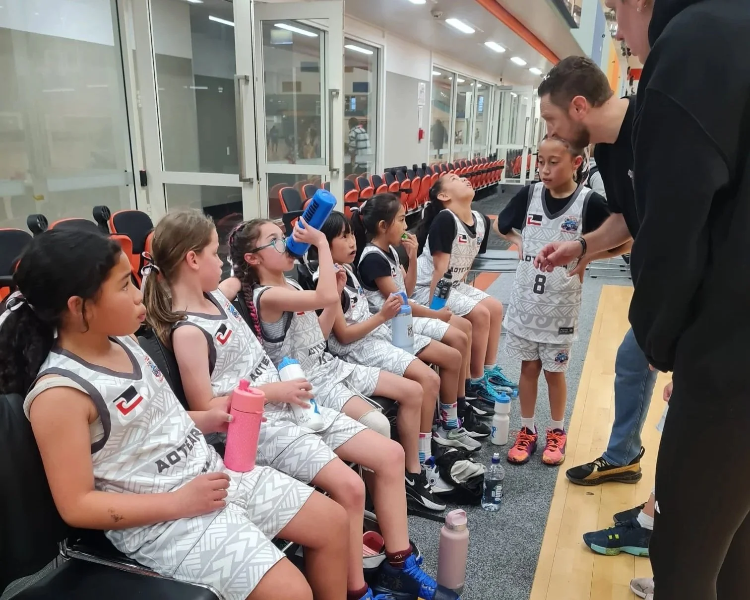 A group of young female basketball players sitting on chairs in a locker room, listening to their coach or trainer who is talking to them. Some are holding water bottles, and one girl is drinking from a blue water bottle.
