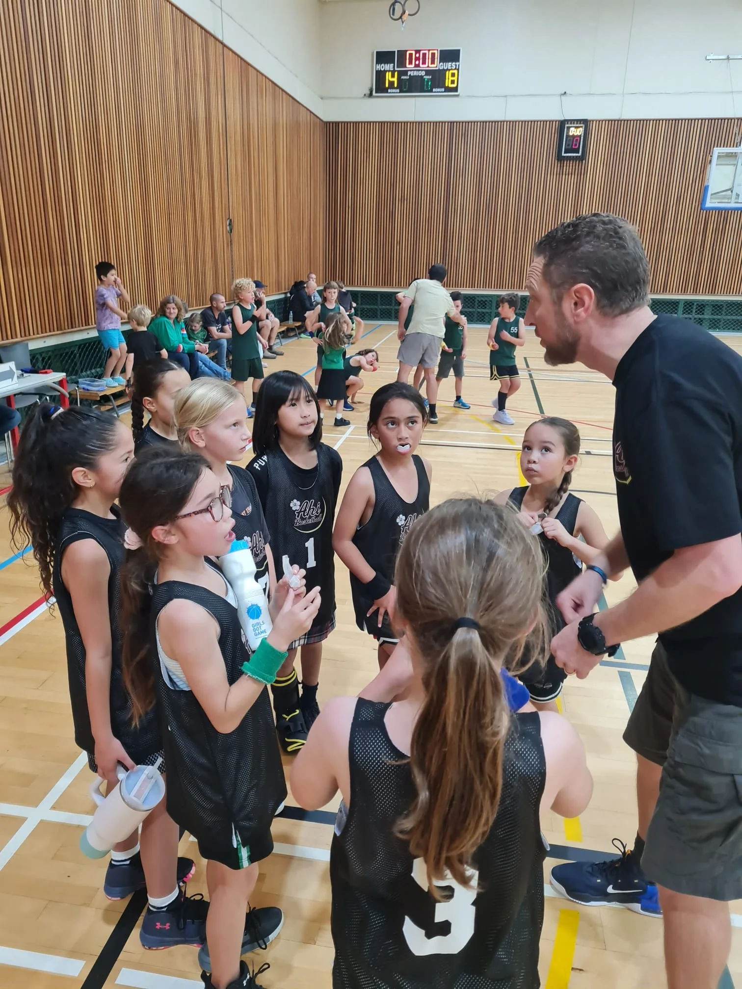 A group of young girls in black basketball uniforms are gathered in a gym talking with a male coach about the game. Other children and adults are in the background, some sitting on benches and others standing on the gym's wooden floor with a scoreboard showing the score and time at the end of a game.