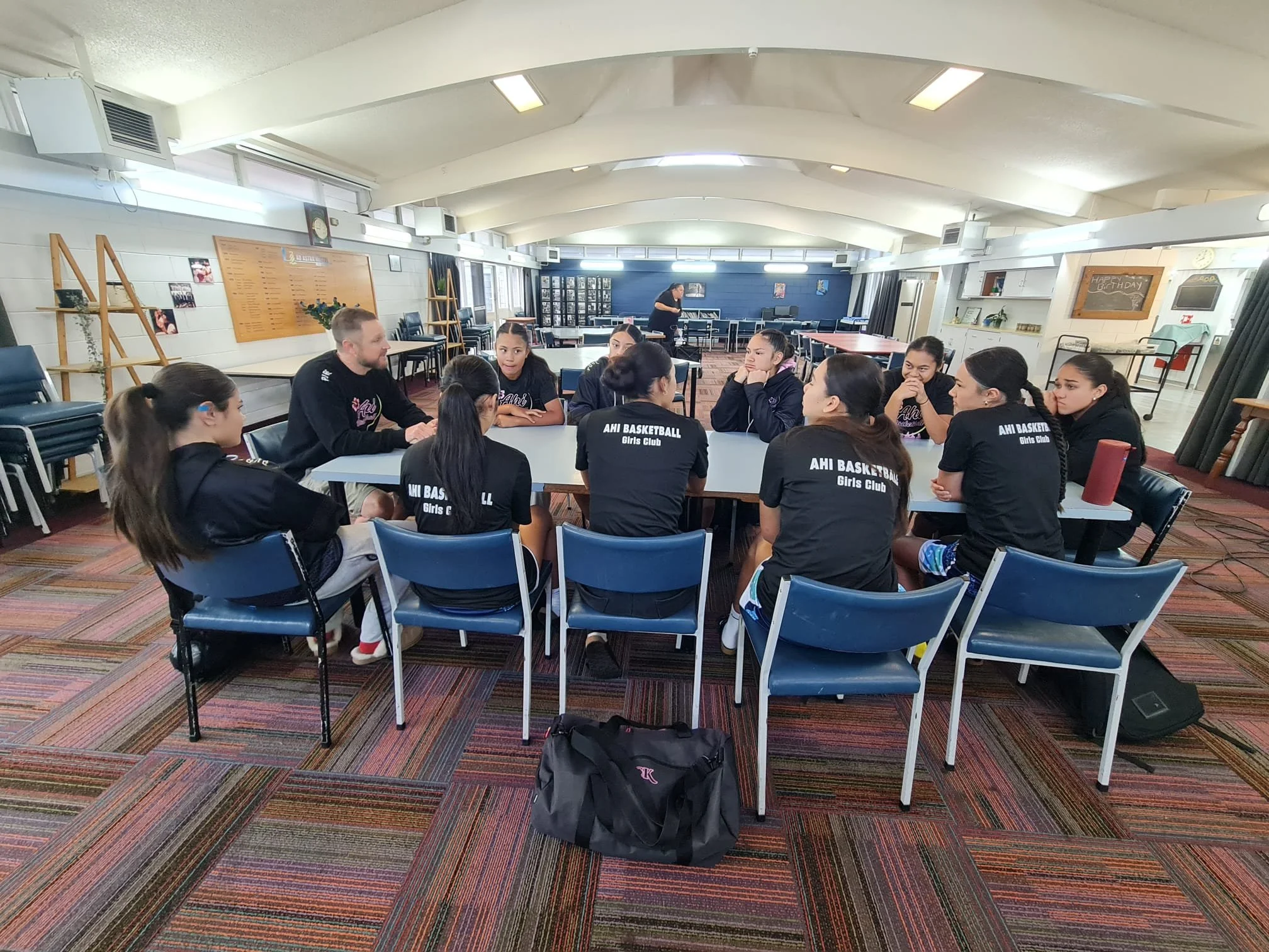 A group of young girls wearing black shirts with the words 'AHI BASKETBALL Girls Club' sitting around a table in a community room with a man, a woman, and a woman in the background.