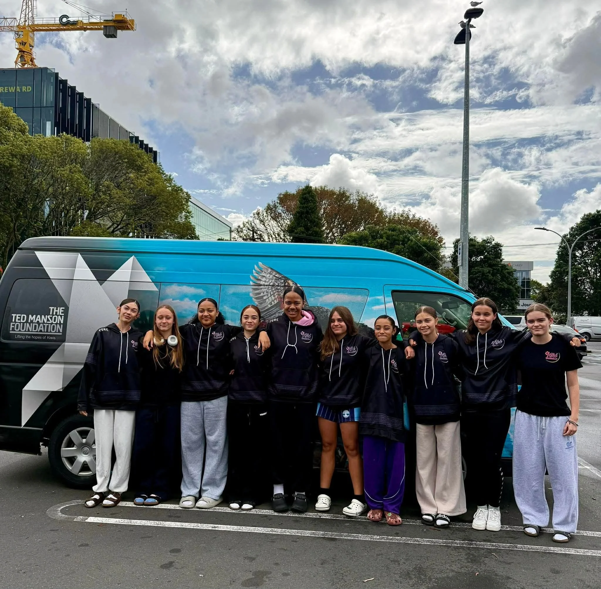 A group of about ten young girls standing together outdoors in front of a van with the logo of The Ted Manson Foundation. The girls are smiling and dressed in matching dark jackets with white lettering. Behind them are trees, a streetlight with pigeons perched on top, and modern buildings under a partly cloudy sky.