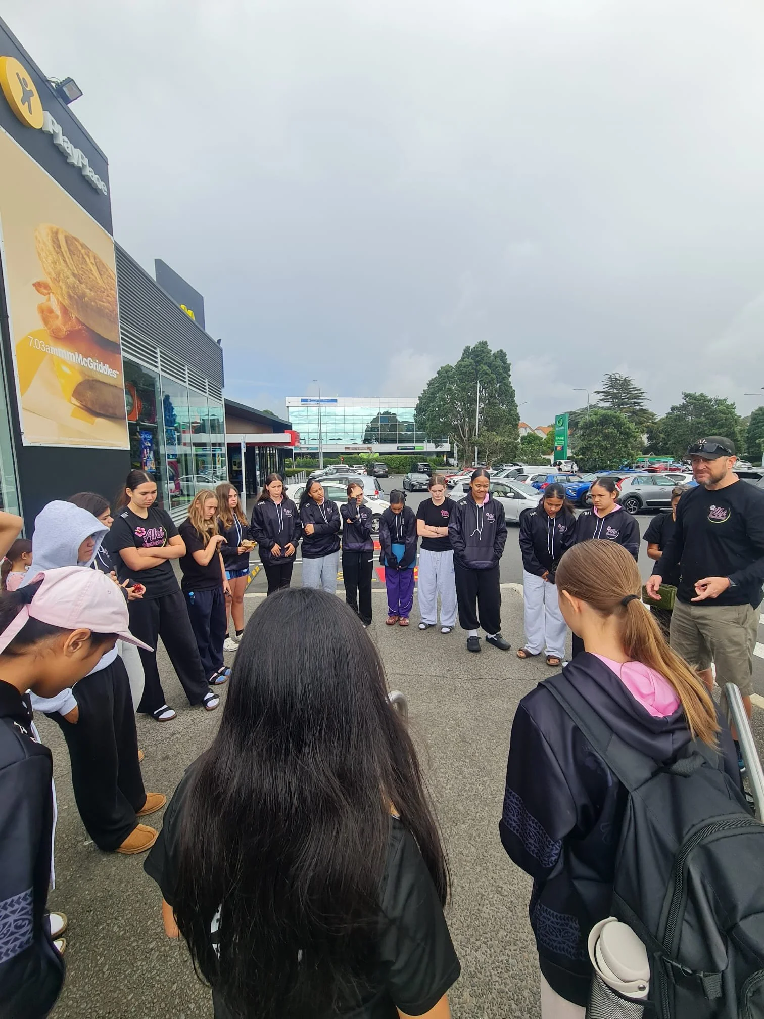 A group of young girls and a man standing in a parking lot, with some people bowing their heads and their hands clasped, at an outdoor gathering near a shopping center or mall.