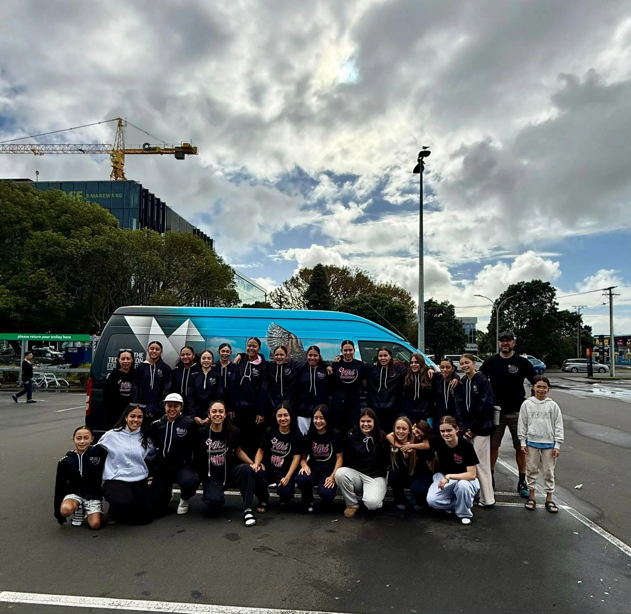 Group of young women and one man gathered outdoors on a parking lot, posing for a photo in front of a blue vehicle, with a cityscape and cloudy sky in the background.