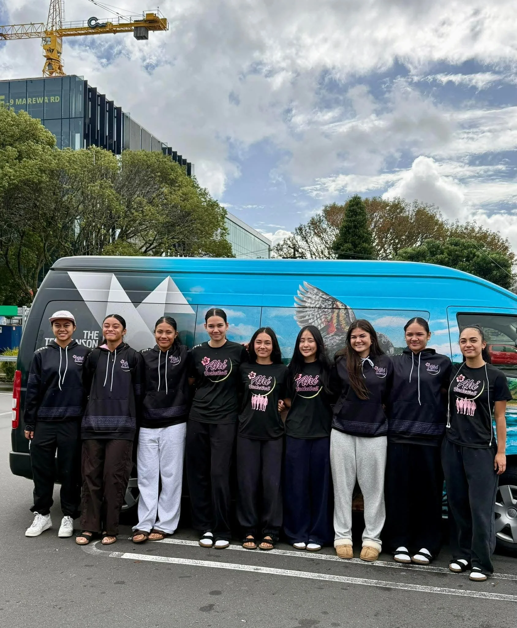A group of young women standing in front of a vehicle with a mural and a building with a construction crane in the background. They are dressed in black hoodies and white or black pants, smiling at the camera.