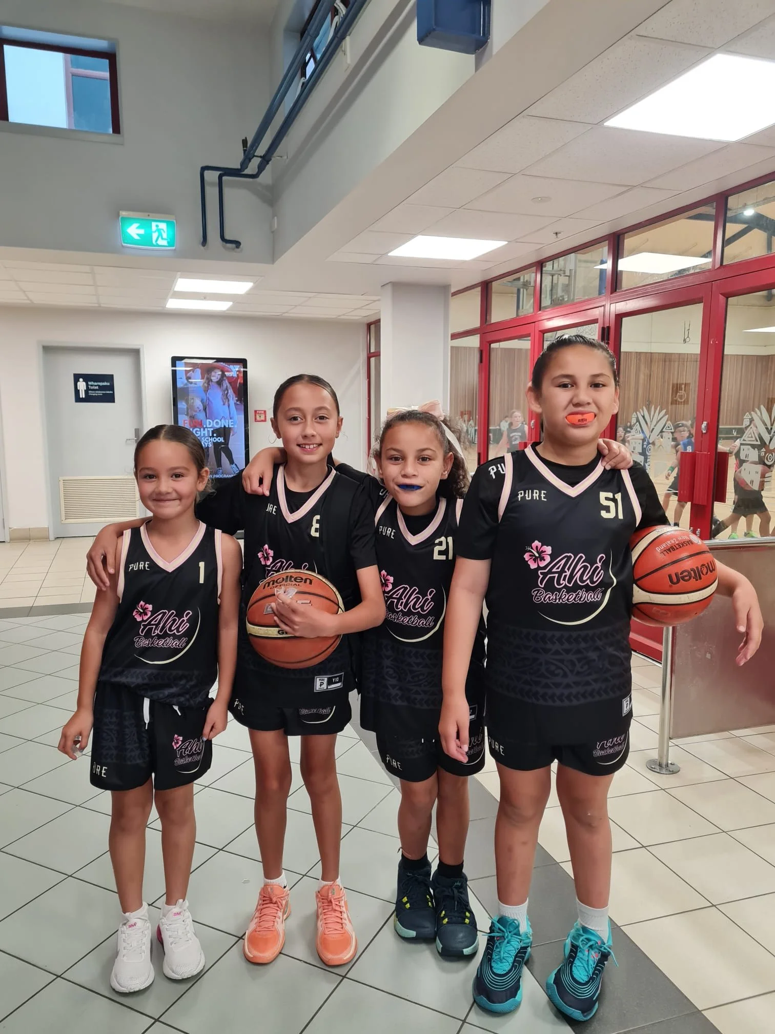 Four young girls in black basketball uniforms with pink and white lettering and numbers, standing inside a building near a red glass door. They are smiling, holding basketballs, and have their arms around each other.