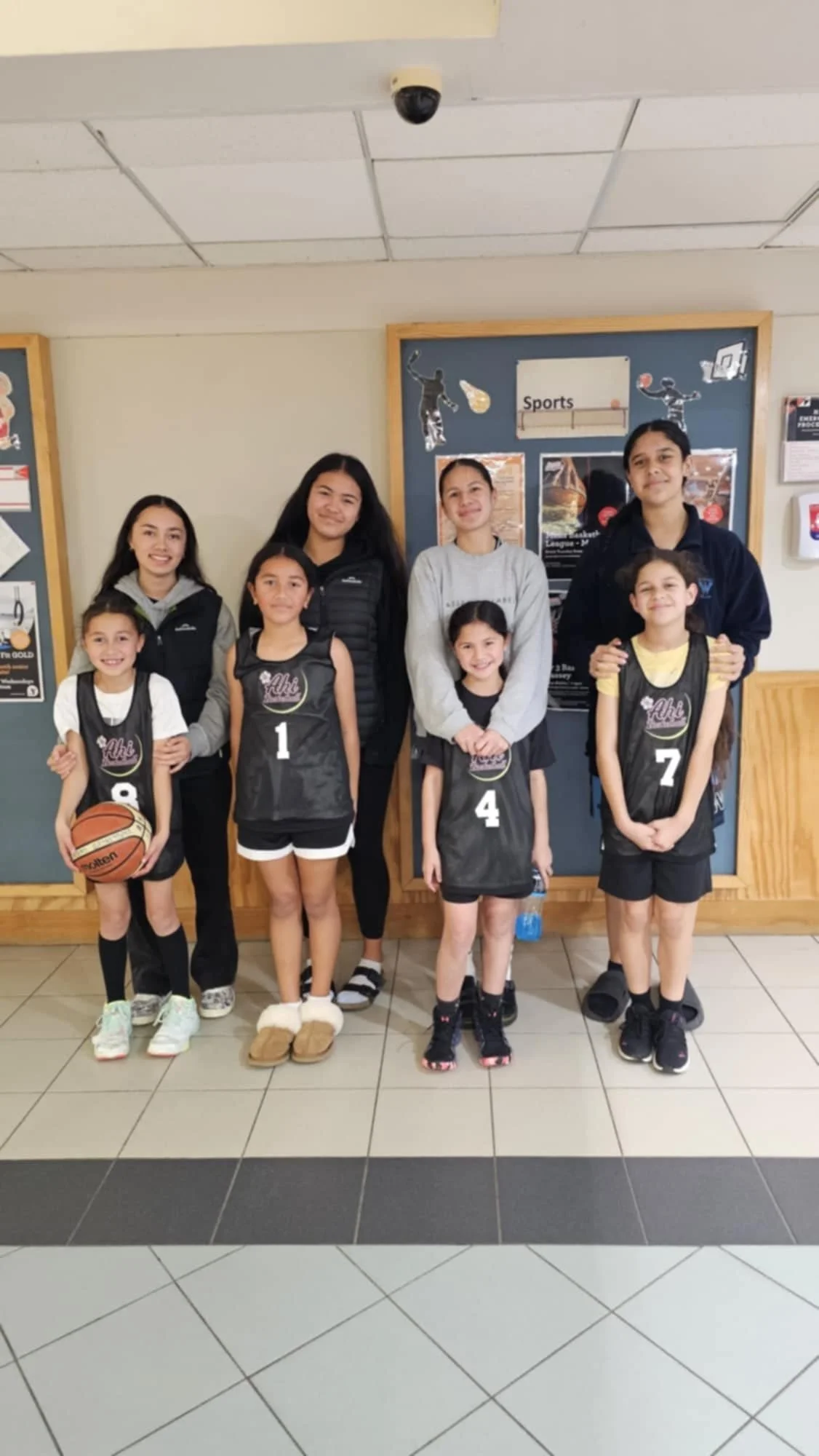 Group photo of young female basketball players in black jerseys with numbers, standing with three teenage girls and one adult woman in casual clothing, inside a building near a sports-themed bulletin board.