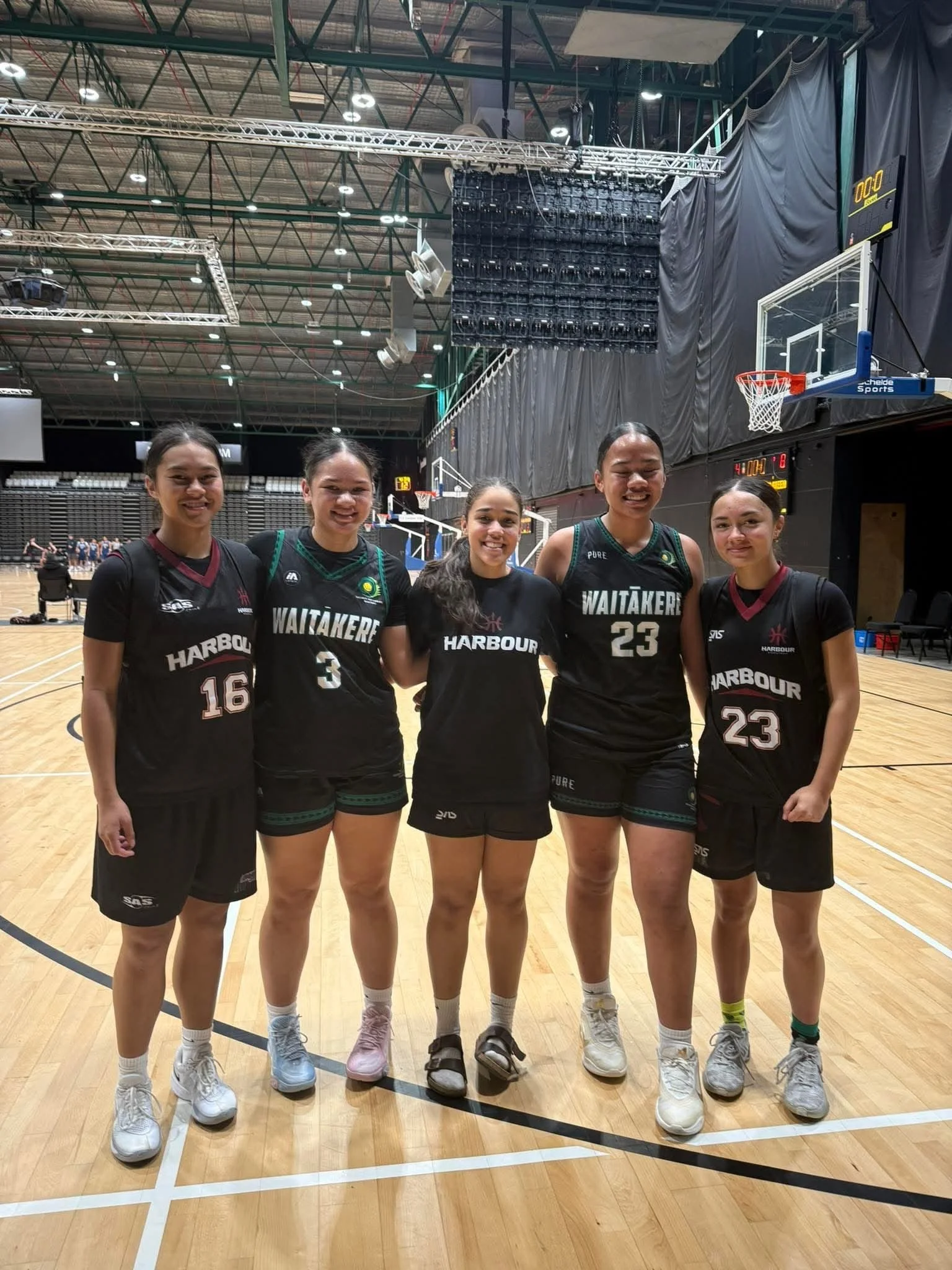 Five young women in black basketball uniforms standing on a basketball court smiling and posing for the photo.