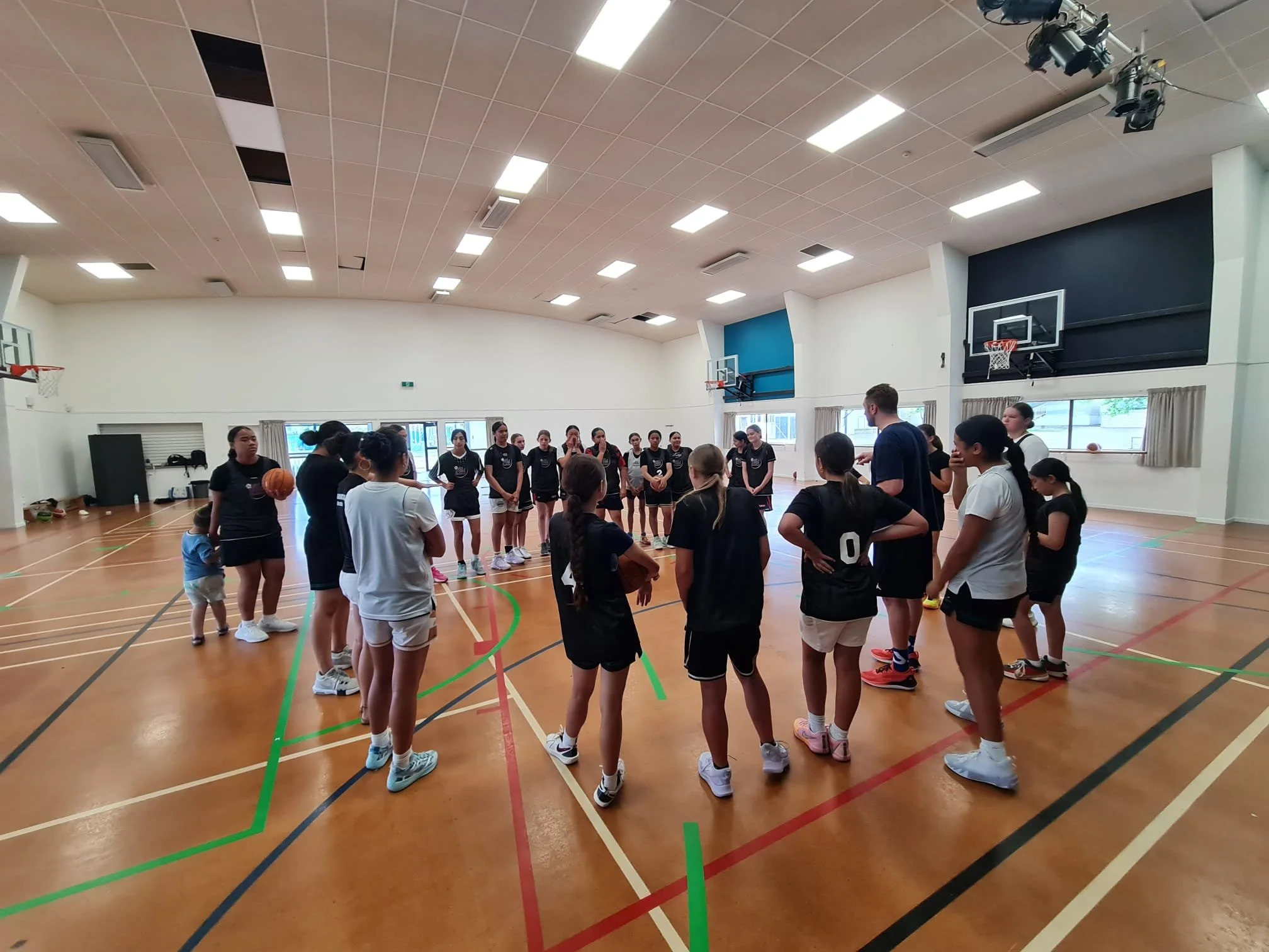 A group of young women in sports uniforms, some holding basketballs, gathered in a gymnasium with a coach or instructor talking to them.