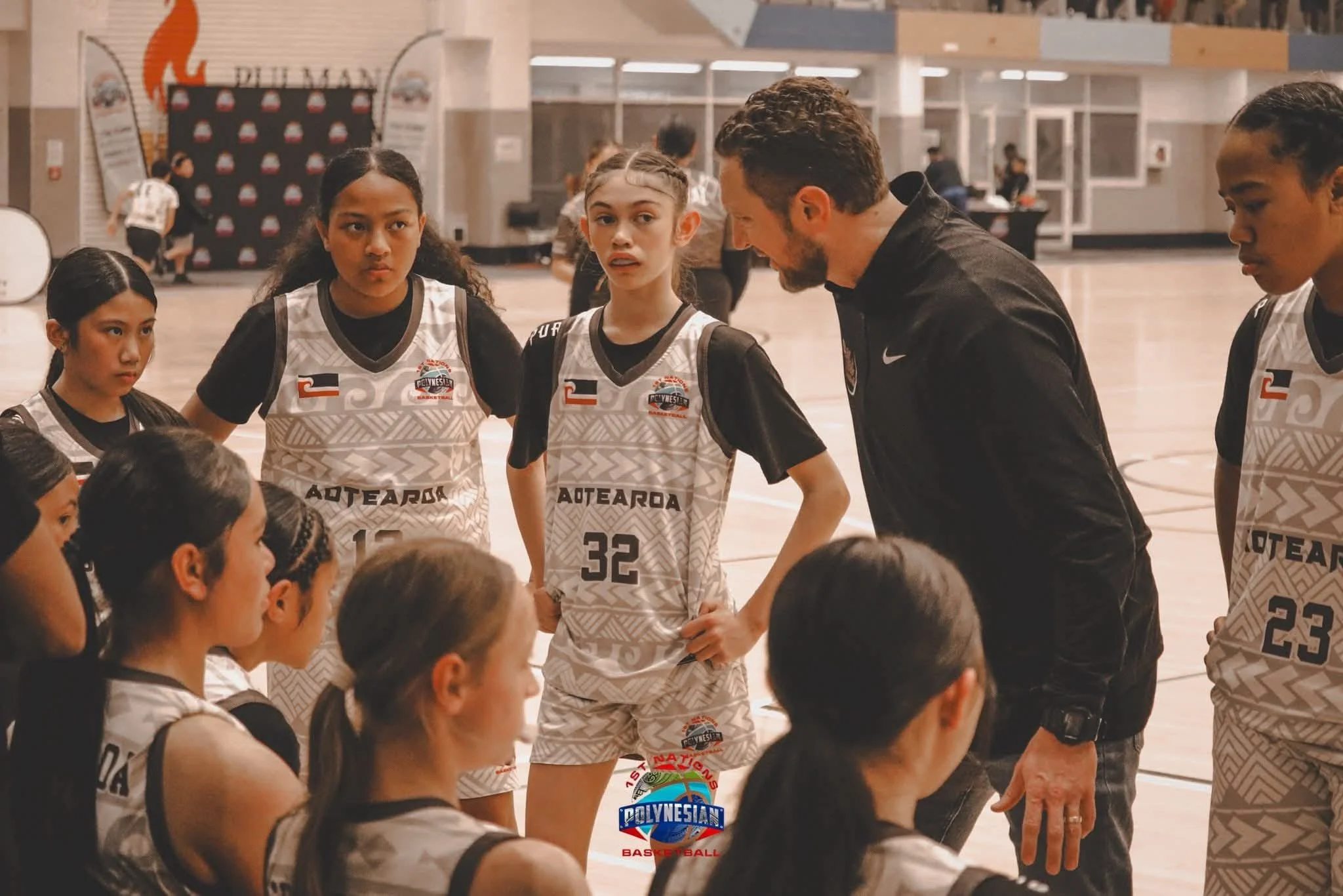 A group of young female basketball players with a coach in a gymnasium during a timeout. The players are wearing Team New Zealand uniforms with numbers, and the coach is speaking to them. Other players are seated on the floor listening.