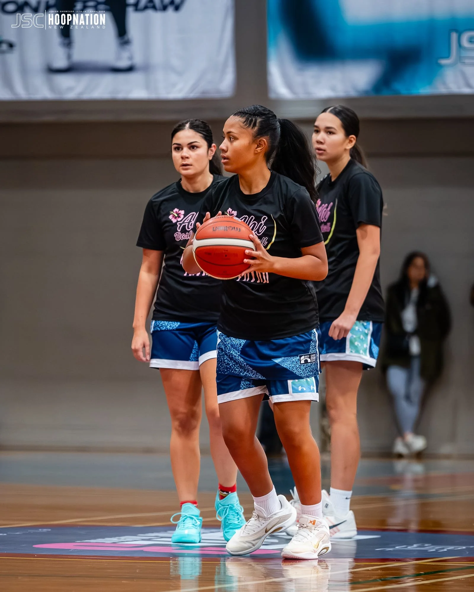 Three female basketball players in black shirts and blue shorts warming up or practicing on an indoor basketball court.