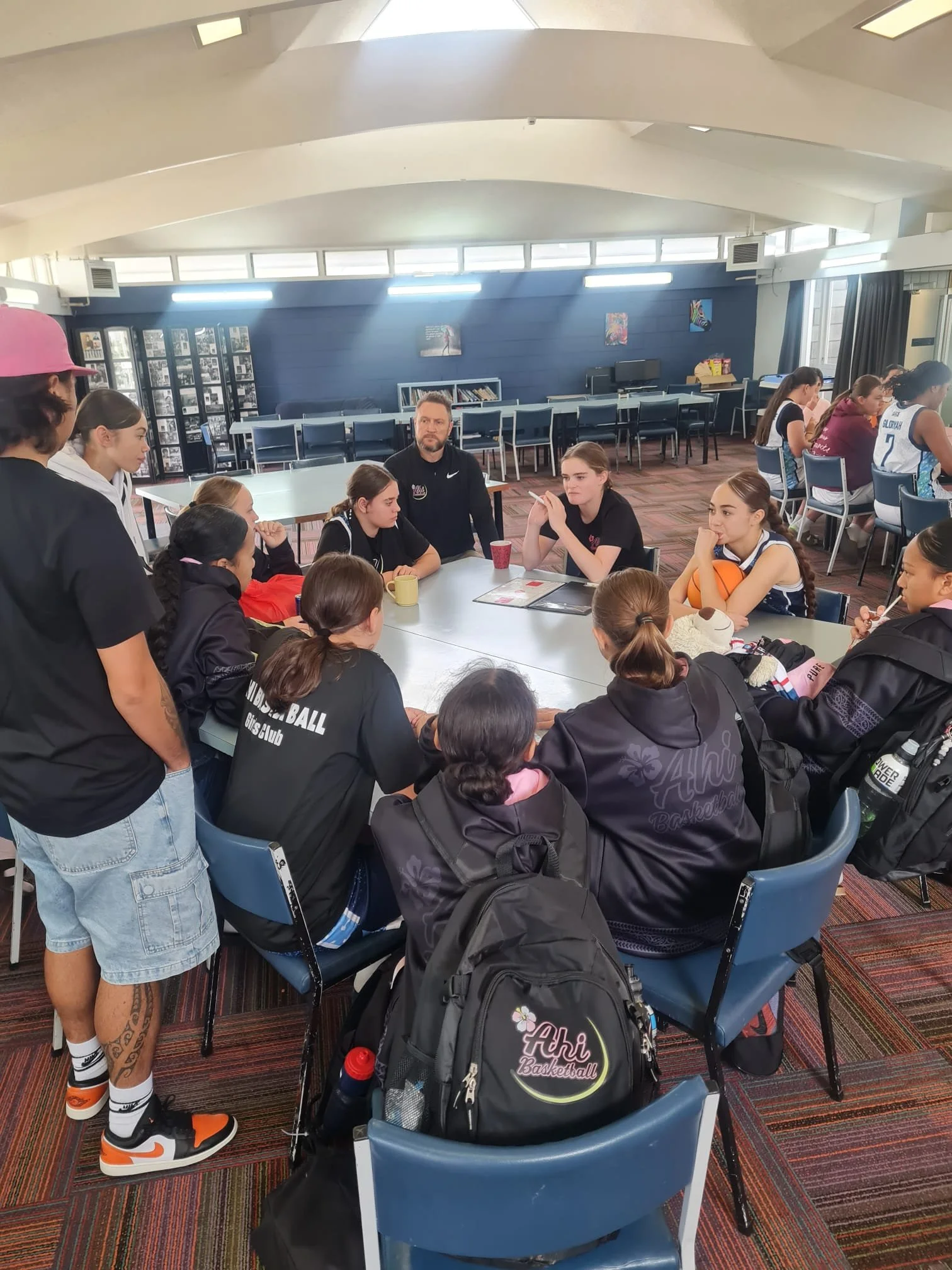 A group of young female basketball players sitting around a table in a room with a dark blue wall, colorful artwork, and a man in black talking with them. Some girls are listening, others are talking or looking at their phones. They are wearing black and white basketball uniforms, some with the name 'Ahi Basketball' on them. The room has a striped carpet floor and a large black basketball backpack with a flower design on the back of one of the chairs.