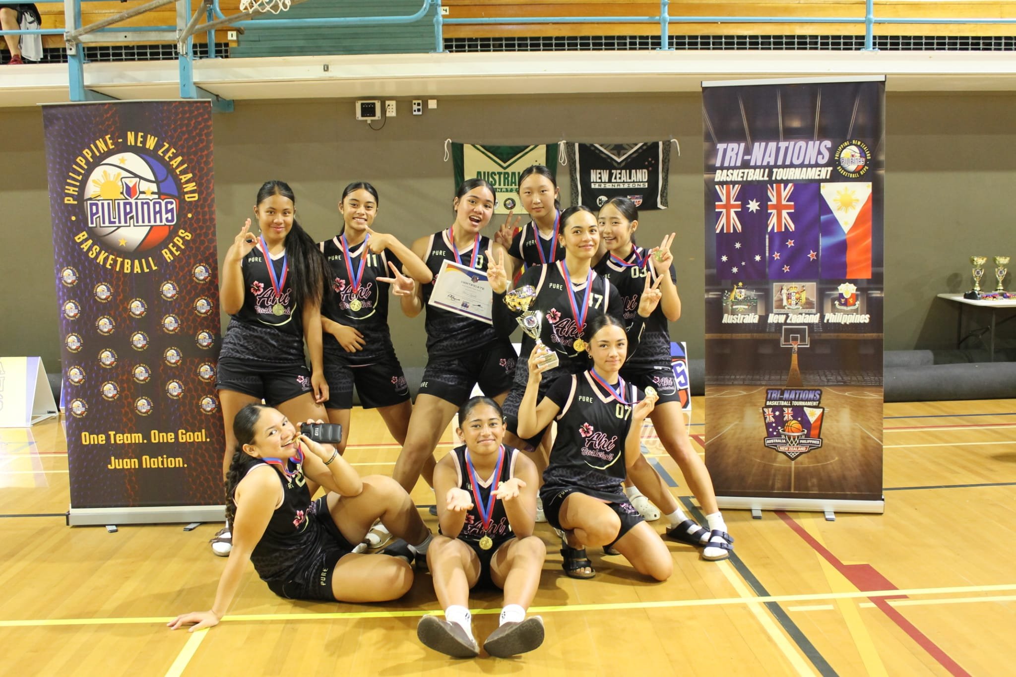 A girls' basketball team in black jerseys celebrating with medals and trophies at a tournament, standing on a gymnasium floor in front of banners that read 'Philippine-New Zealand Basketball Reps' and 'Tri-Nations Basketball Tournament.'
